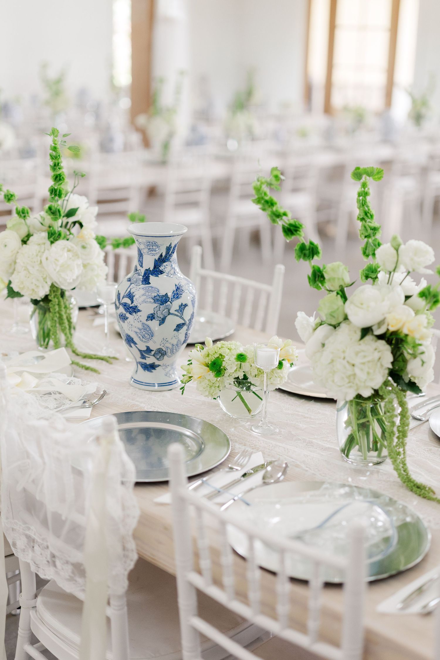 Elegant wedding reception table with white floral arrangements, blue and white chinoiserie vase, and white chiavari chairs.