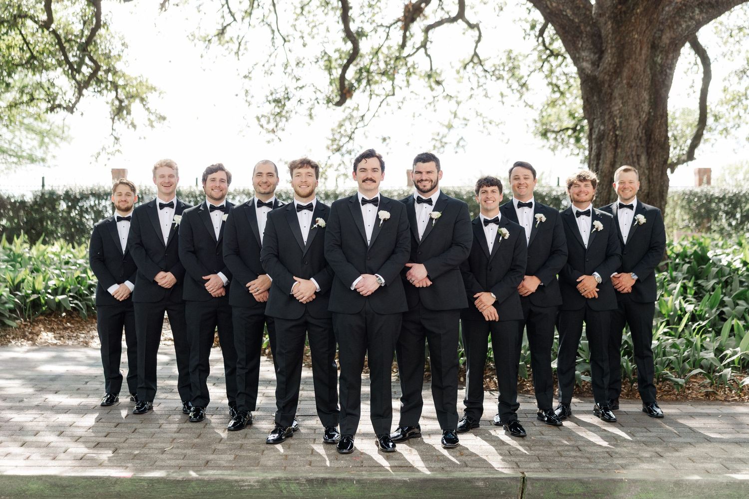 Groom and groomsmen pose in matching black tuxedos and bow ties outdoors at wedding.