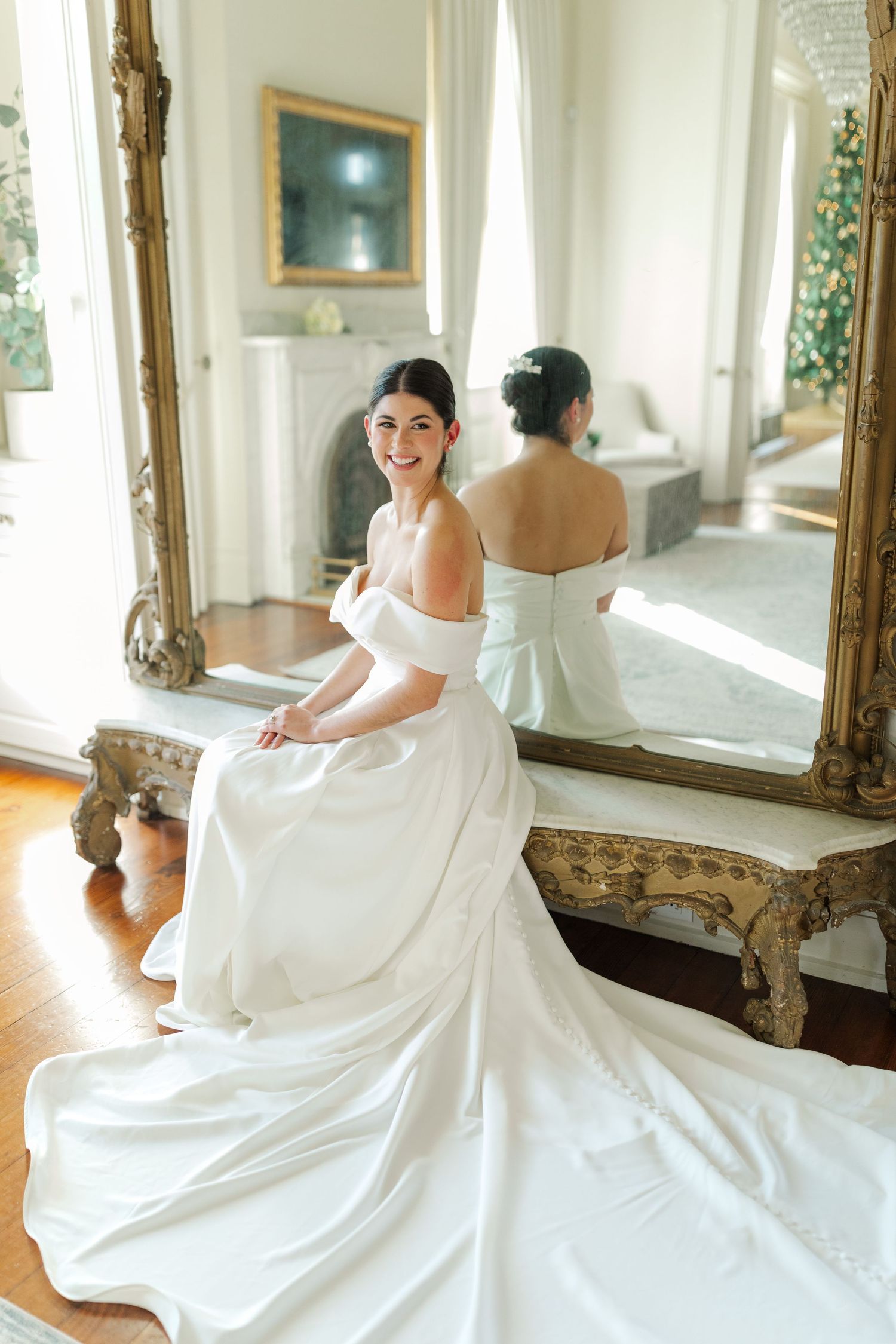 A bride in a white off-shoulder gown sits by an elegant gilded mirror in a sun-lit room.