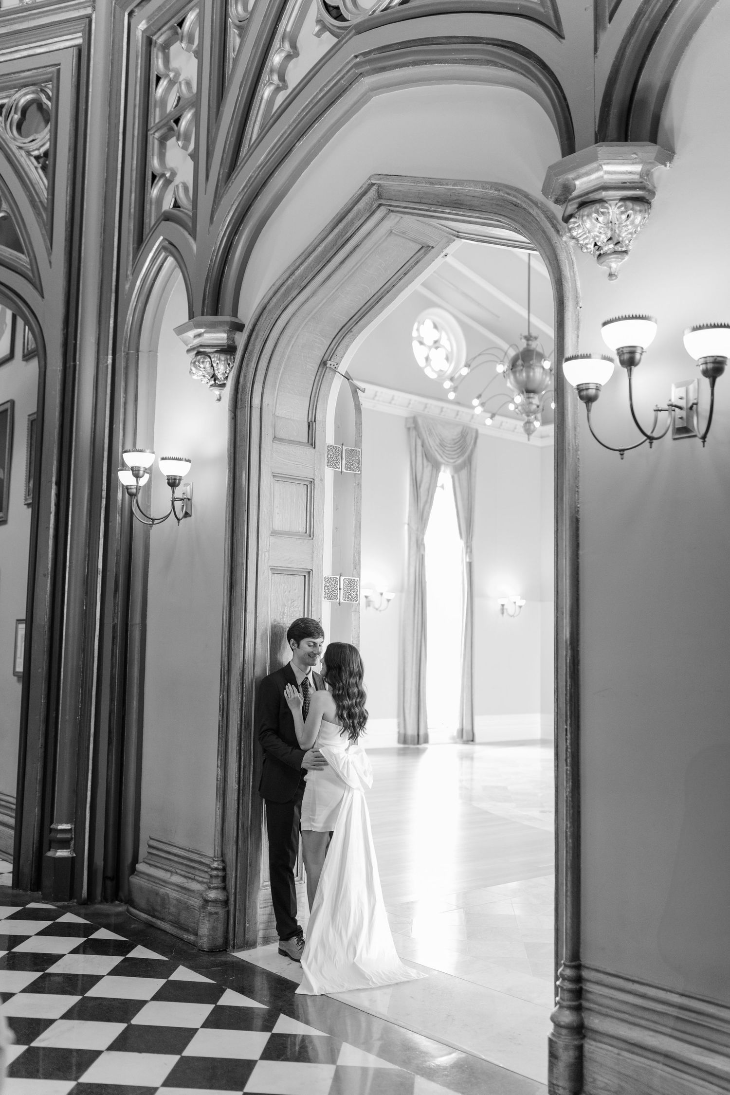 A romantic black and white wedding photo captured in an elegant hallway with ornate arches and checkerboard floors.