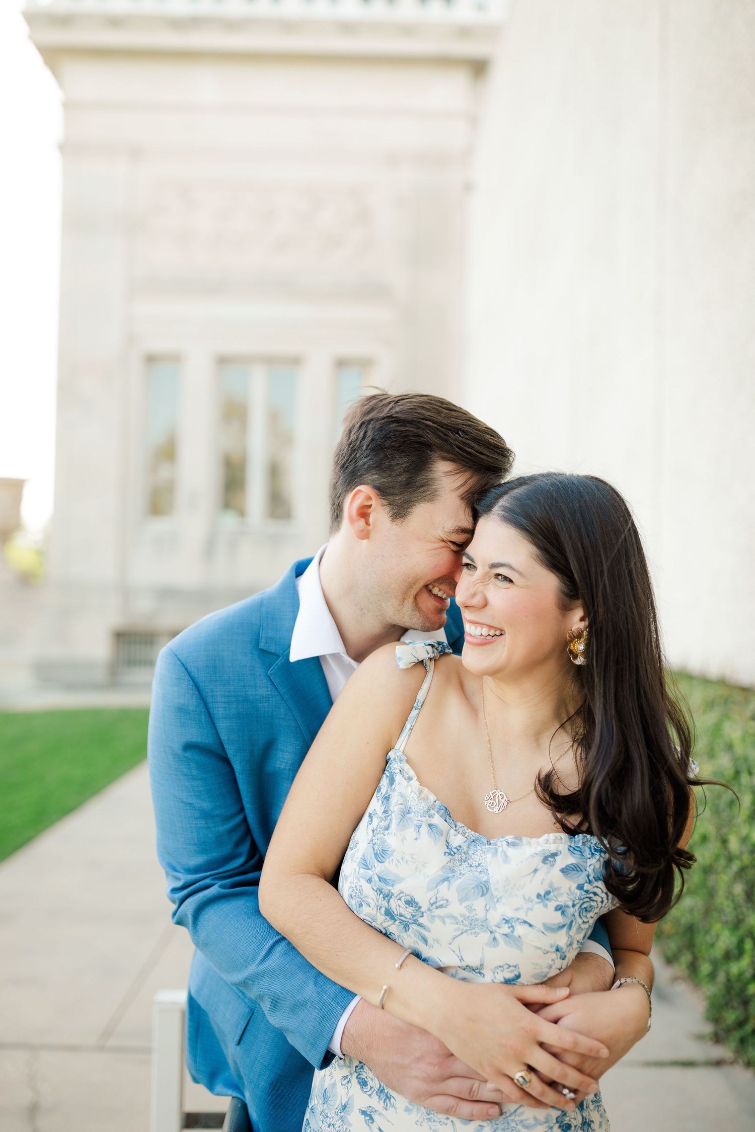 A couple shares a joyful embrace while wearing blue and floral outfits against a light stone building exterior.