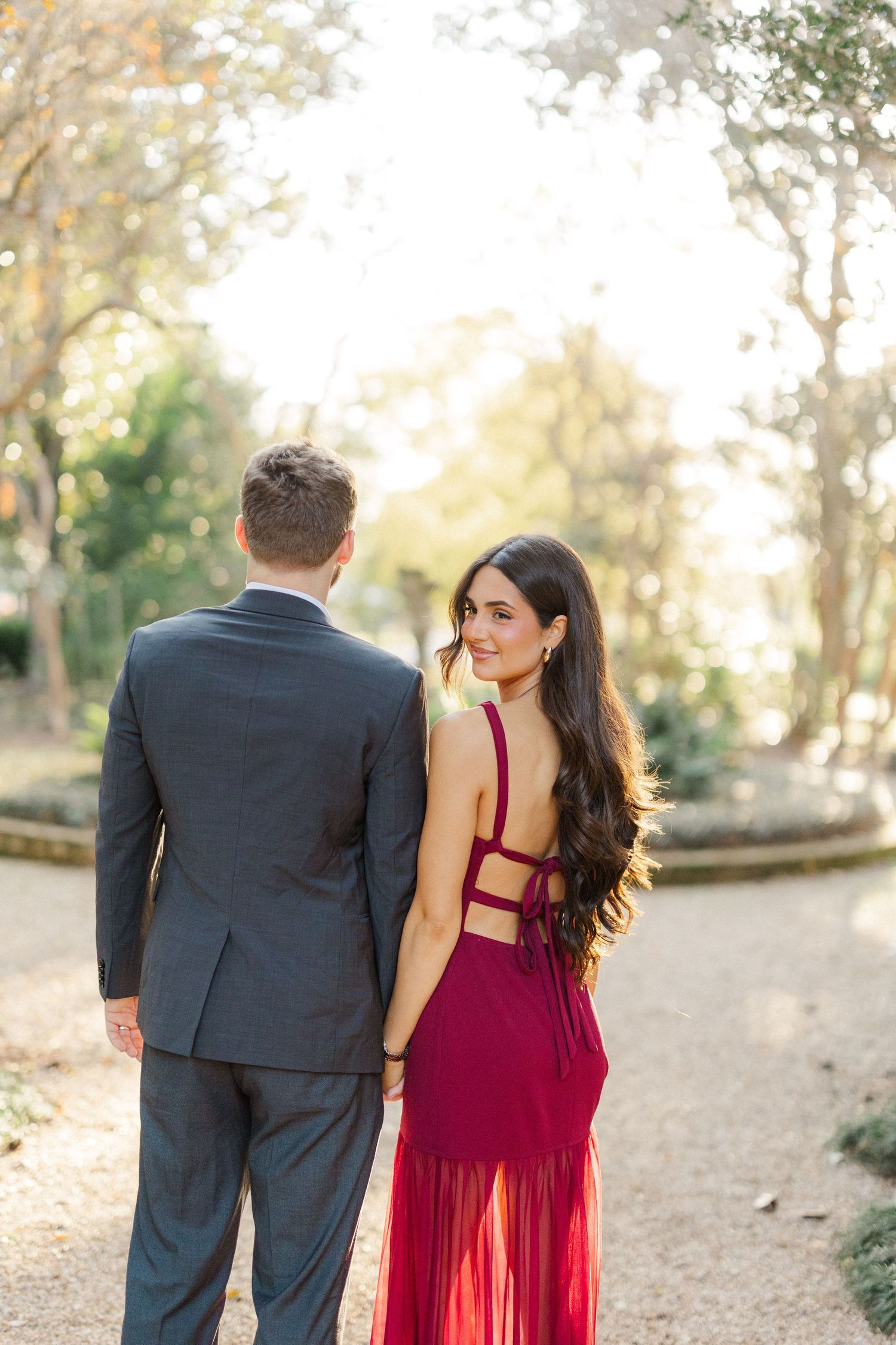 Couple in formal attire poses on garden path with her burgundy dress featuring strappy back detail.