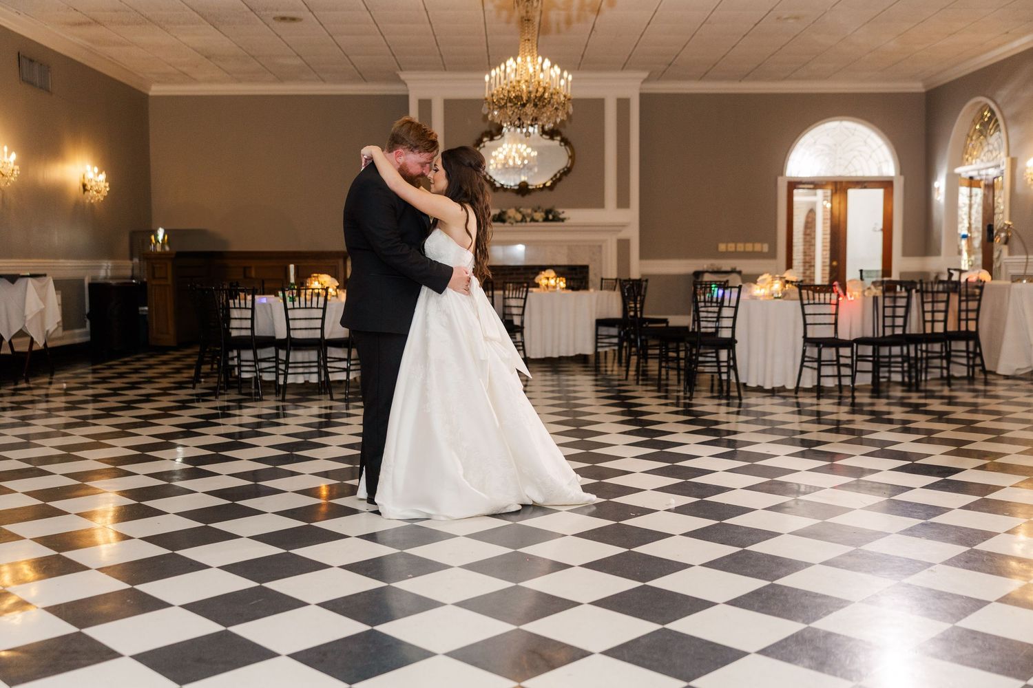 A couple shares their first dance on a black and white checkered floor under an elegant chandelier.