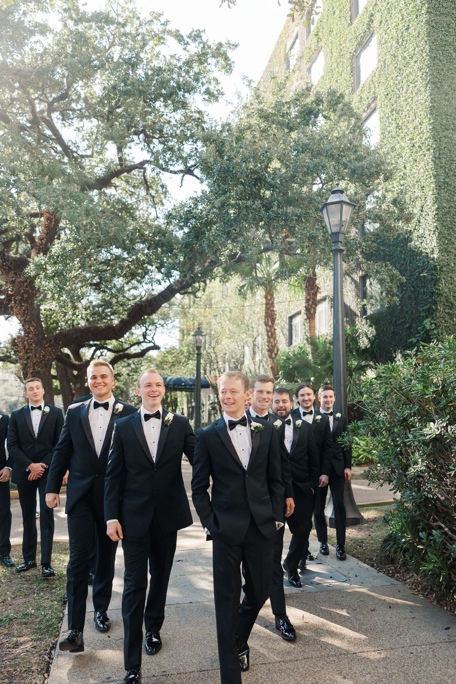 Groomsmen in black tuxedos walk together on a tree-lined path during a wedding celebration.