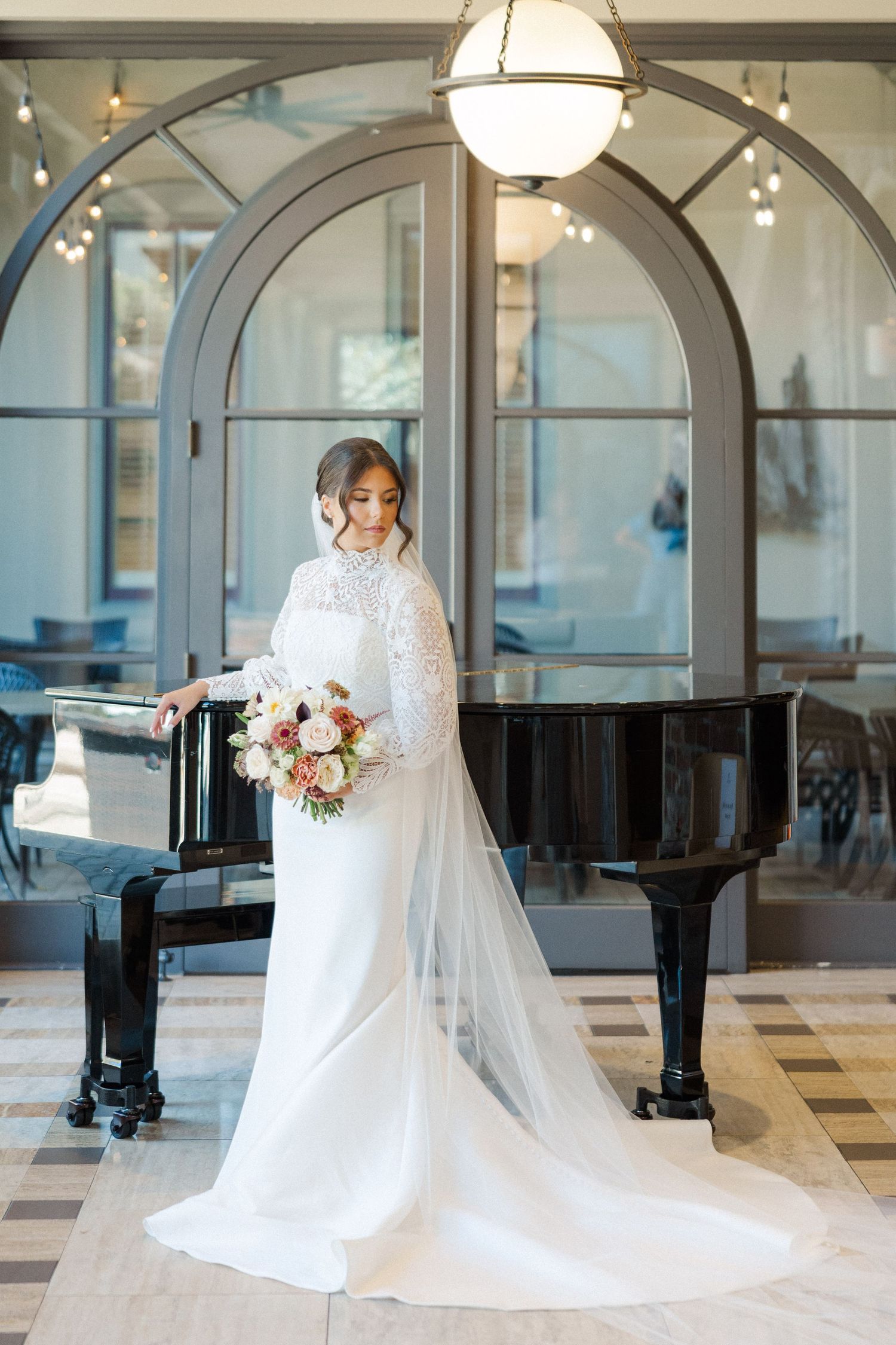 Elegant bridal portrait in white lace gown with long train and veil standing beside grand piano in sunlit venue.