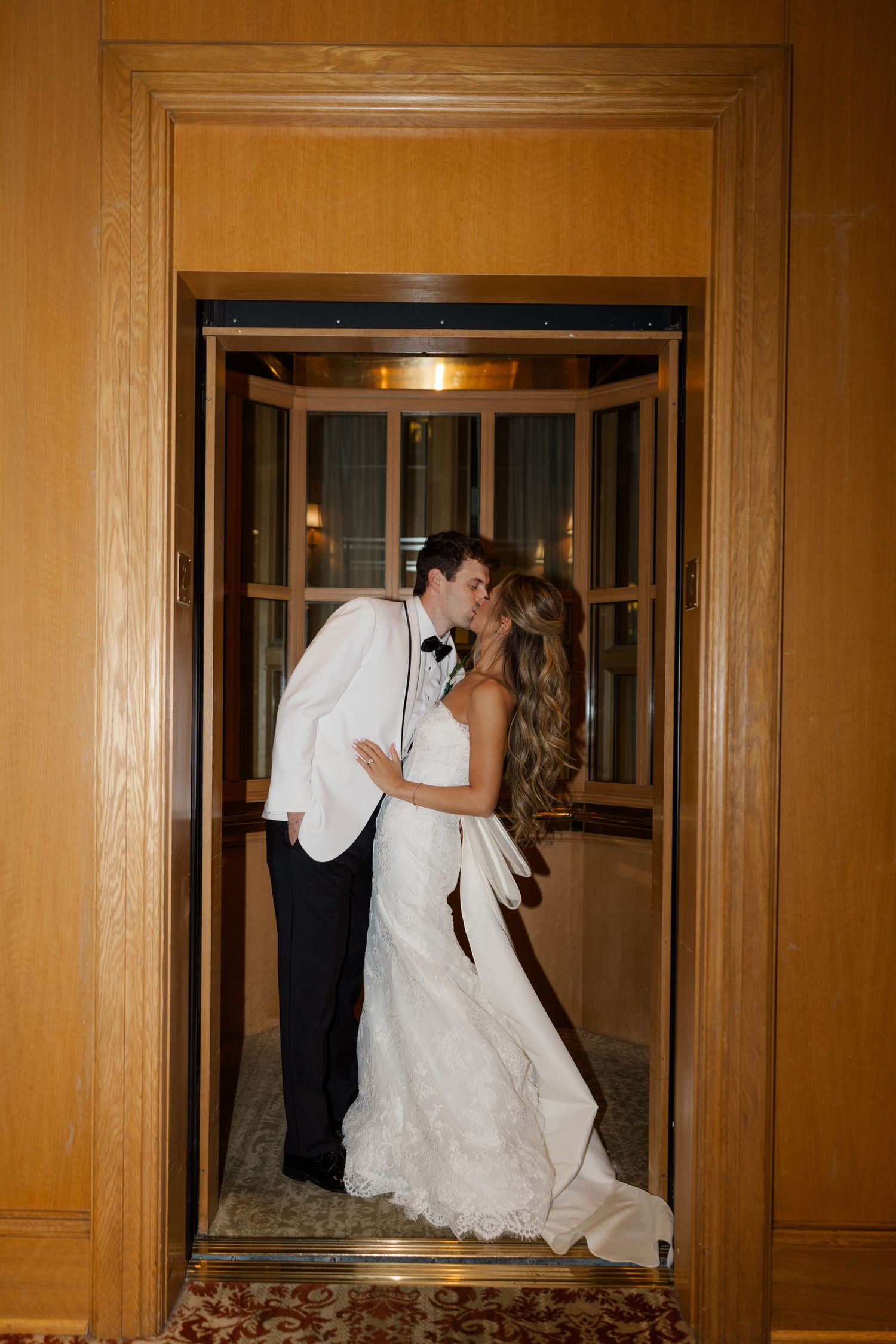 A couple shares a romantic kiss in a golden-lit elevator doorway at their wedding.