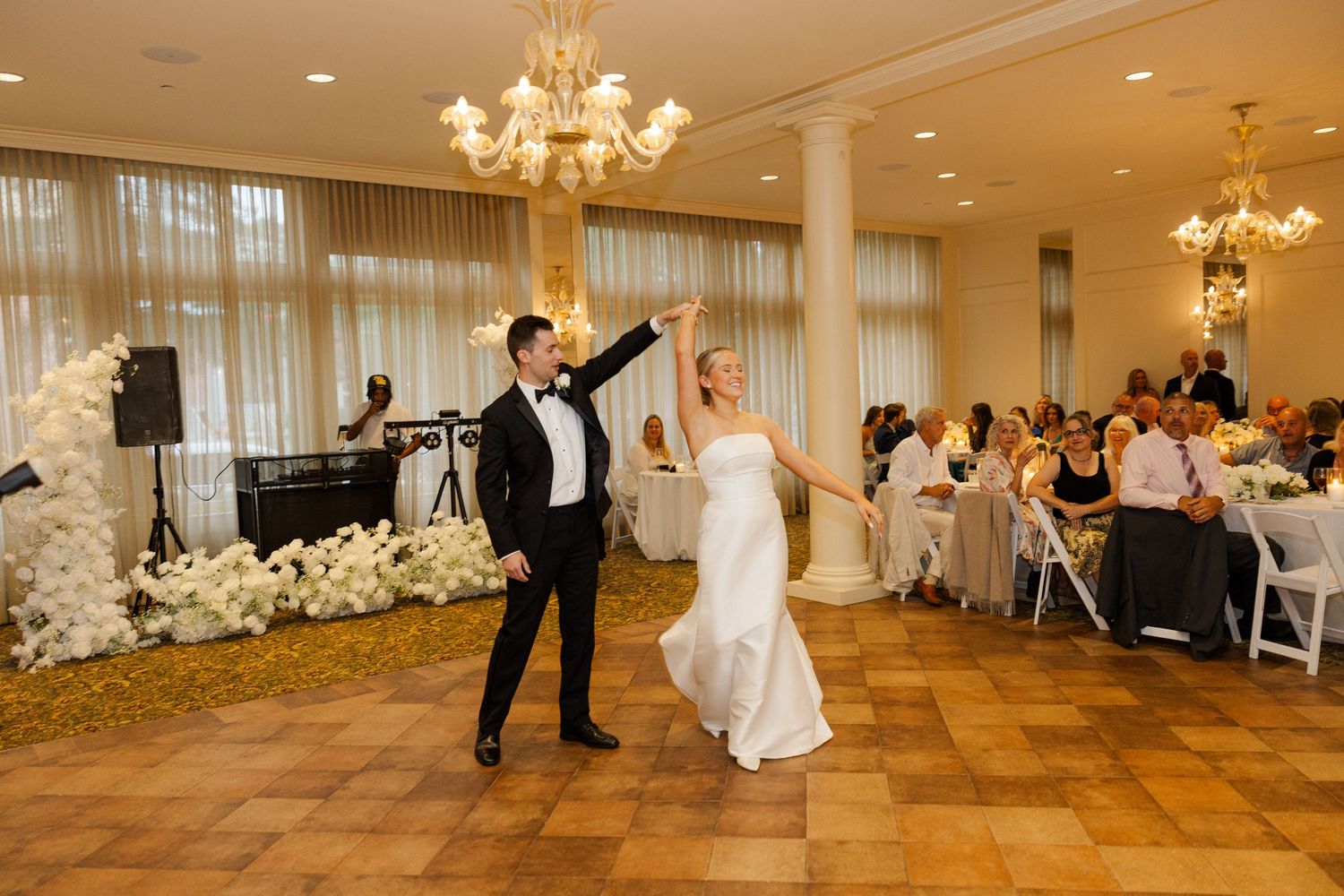 Wedding reception ballroom with chandelier lighting and couples dancing on wooden floor.