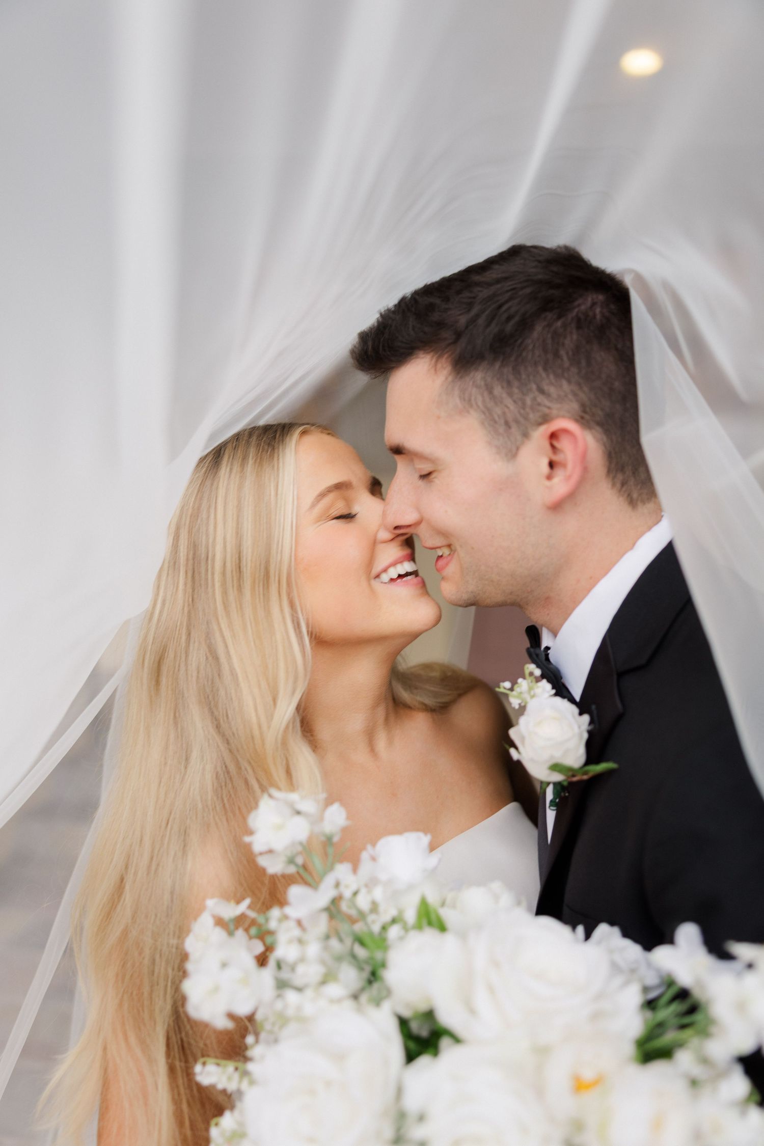 Romantic wedding couple shares intimate moment with white floral bouquet and sheer veil in elegant portrait series.