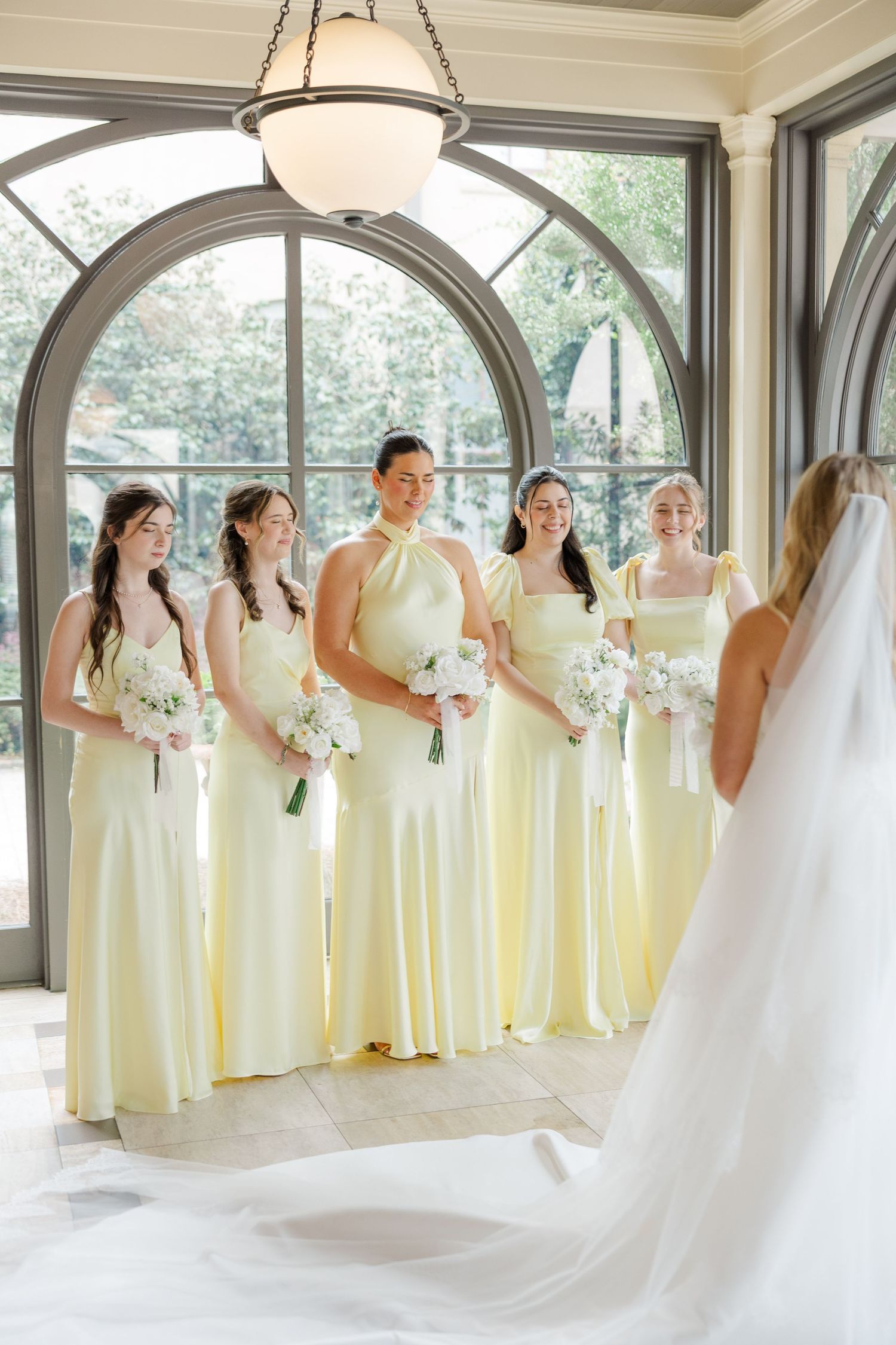 Wedding party in pale yellow dresses and white gown posed in an elegant indoor venue with arched windows.