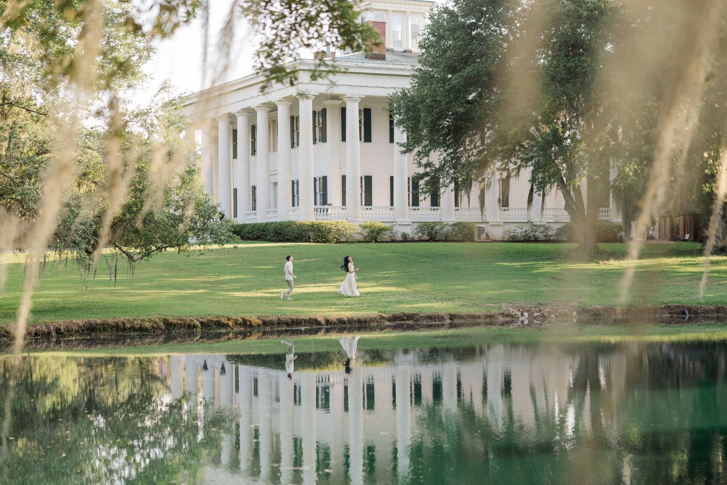 Couple walking on lush green lawn beside a reflective pond in front of a grand white columned Southern mansion.