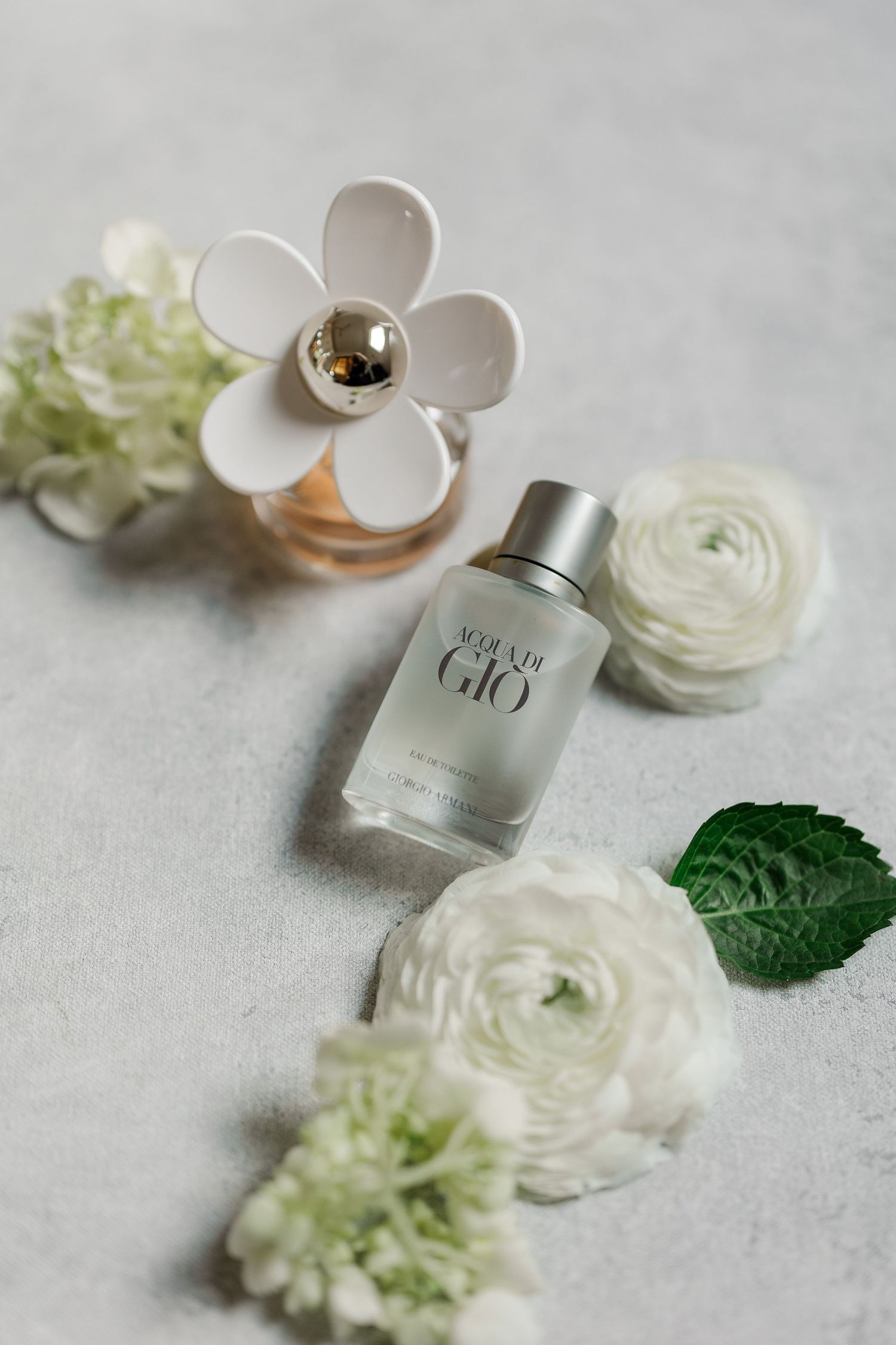 Acqua di Gio cologne bottle surrounded by white ranunculus flowers and a floral-topped perfume bottle on a light gray surface.