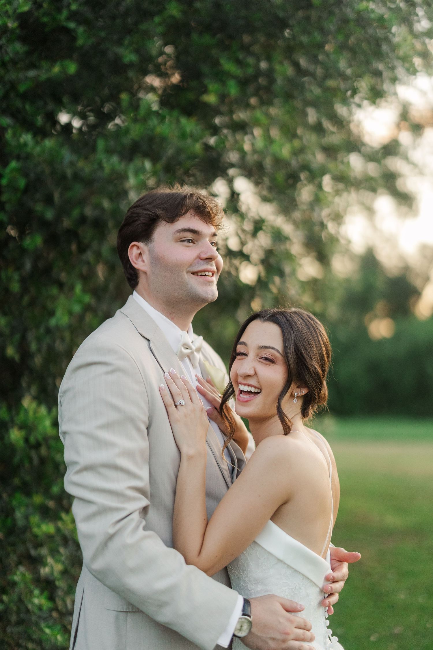 Laughing bride and groom embrace outdoors at golden hour near lush green trees in wedding attire.