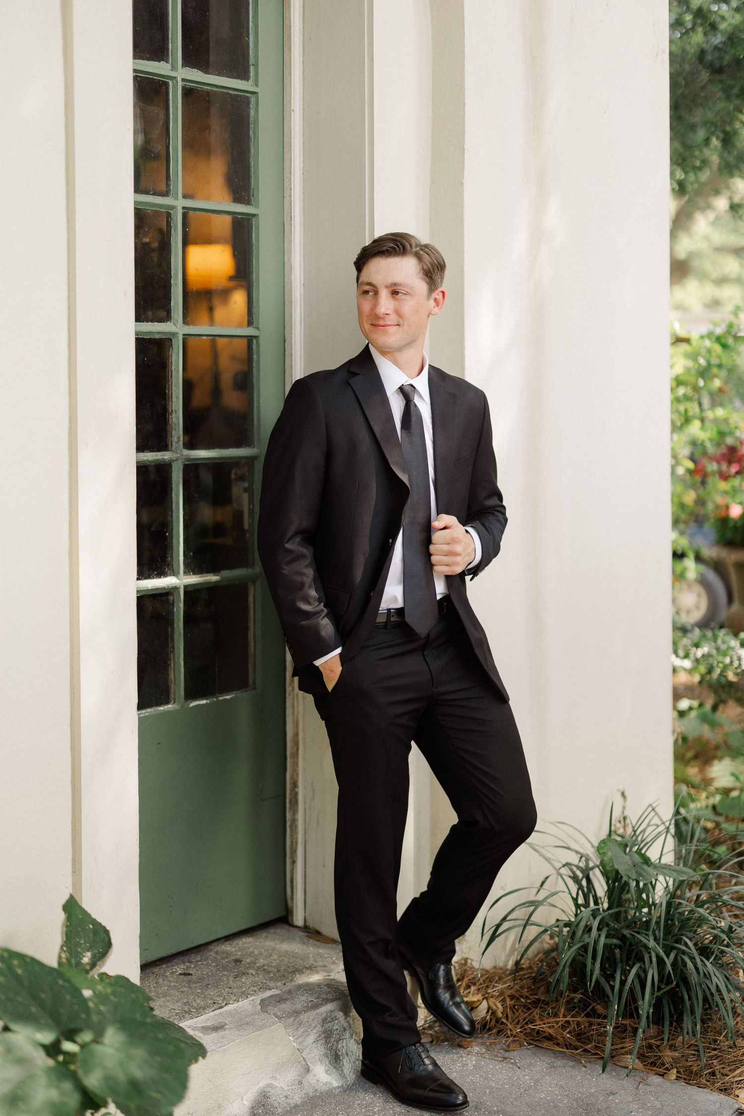 Dapper groom in a black tuxedo poses confidently beside a green French door at an elegant wedding venue.