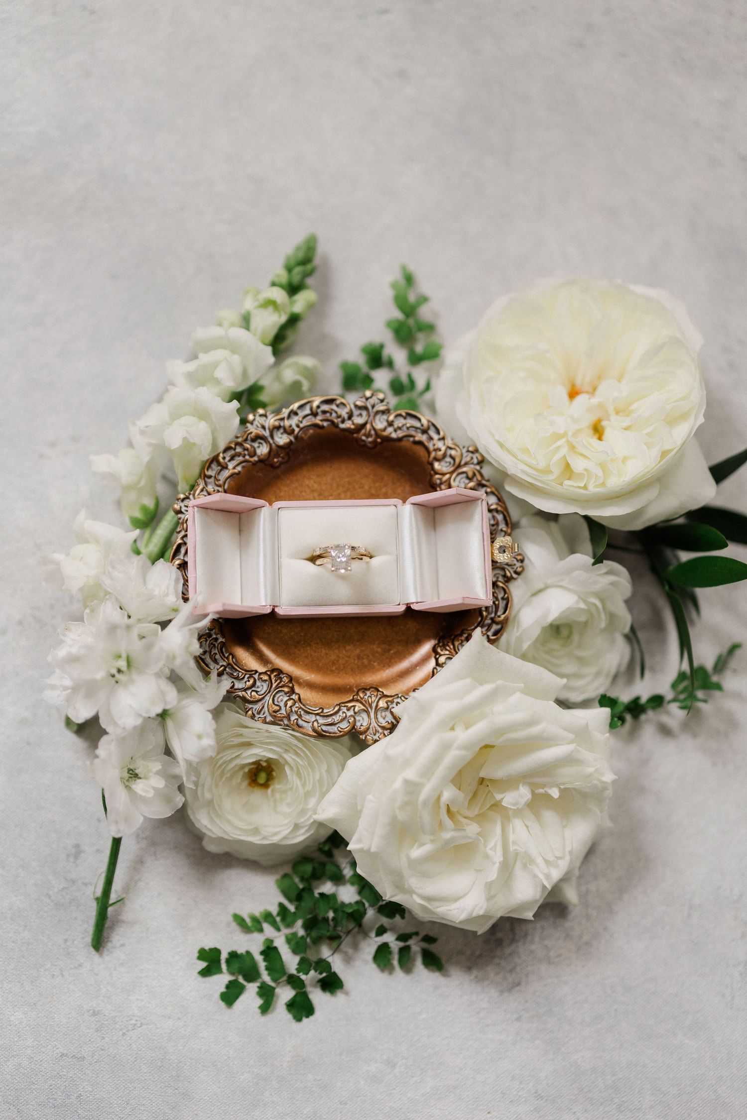 Gold engagement ring with marquise diamond in a pink velvet box surrounded by white roses and ranunculus on a gray surface.