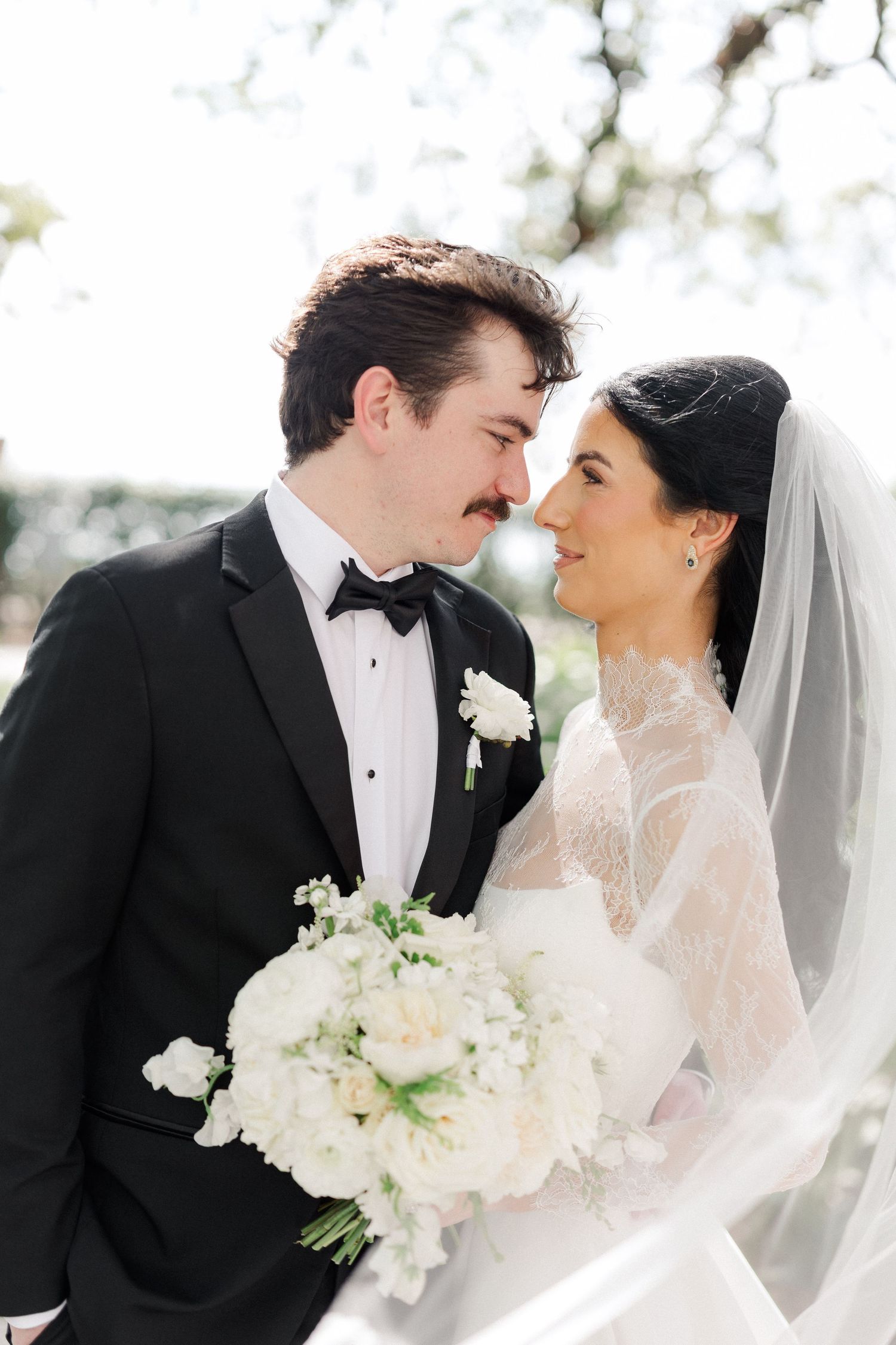 Bride and groom touch foreheads romantically, groom holding white floral bouquet, veil flowing in breeze.