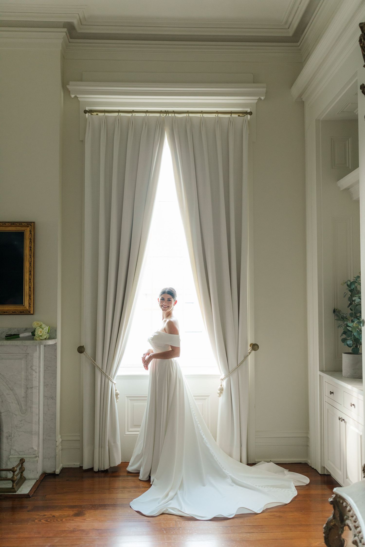 A flowing white wedding gown stands elegantly framed by long curtains in a classic interior with hardwood floors.