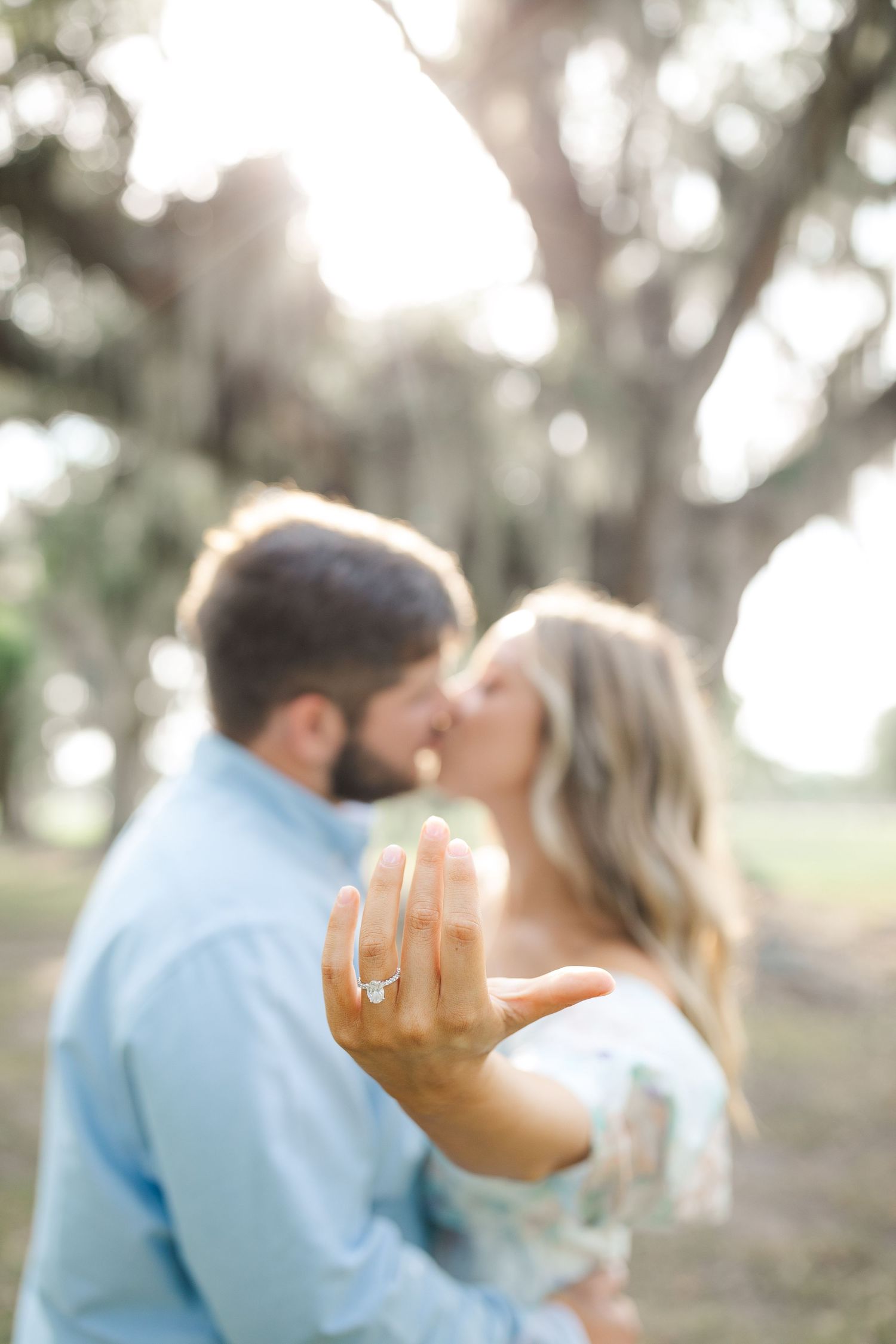 An engaged couple kisses while showing off their engagement ring in soft natural lighting.