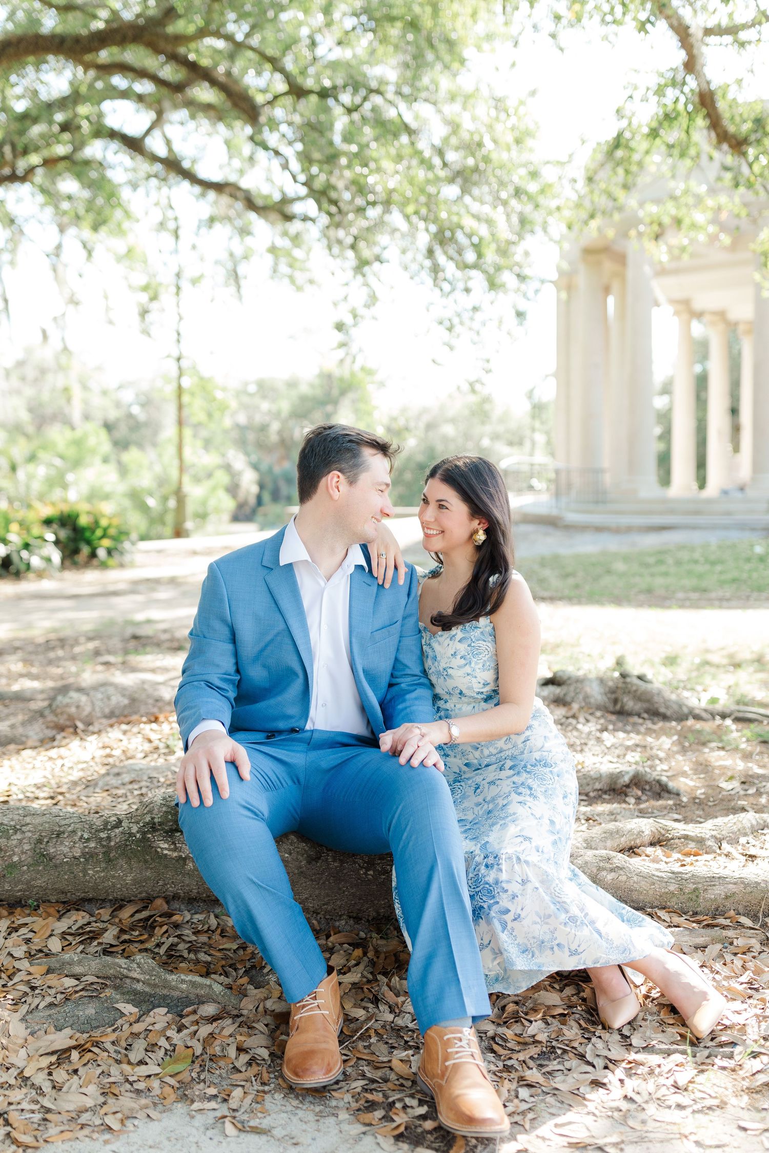 A couple in blue and white formal attire sits together on a tree root near a columned structure in a sunlit garden.