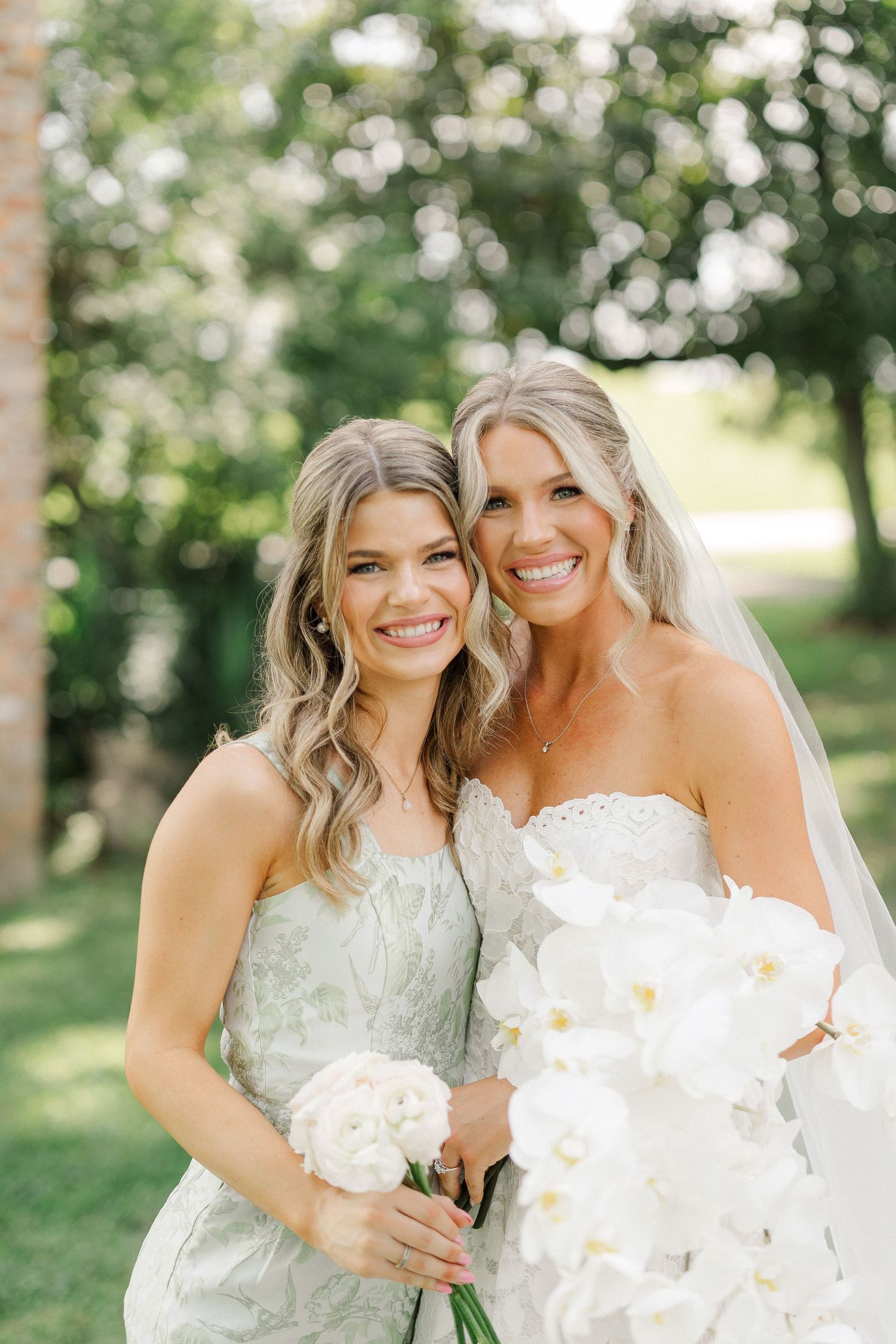 Bride in white strapless gown with veil holding orchid bouquet poses with bridesmaid in sage dress outdoors.