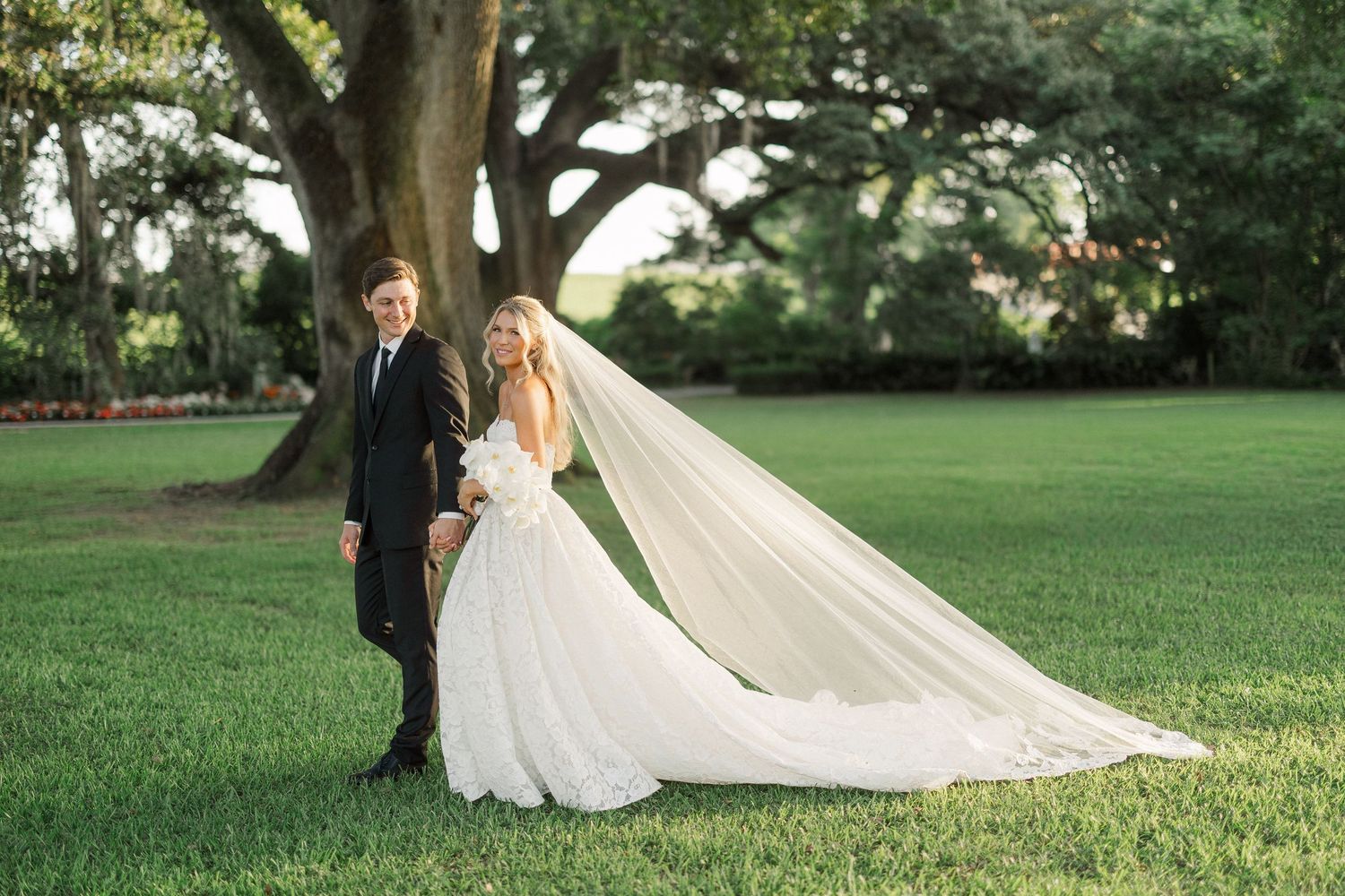 Bride and groom share romantic moment on manicured lawn with dramatic cathedral veil flowing behind large oak.
