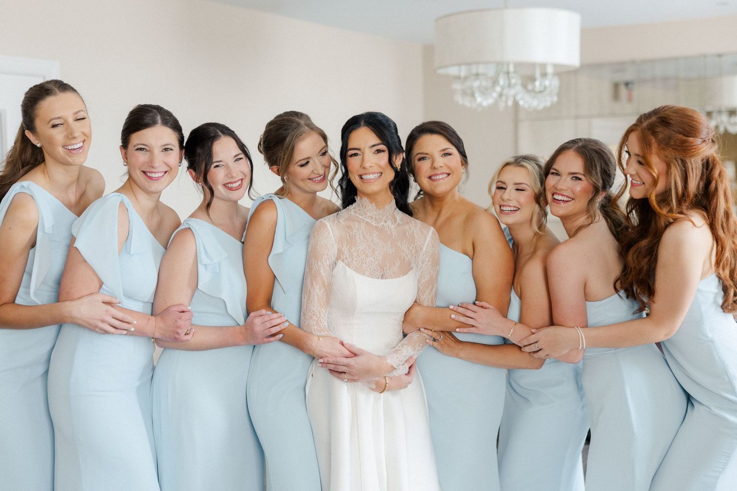 Bride in white lace gown surrounded by seven bridesmaids in light blue dresses, smiling together indoors.
