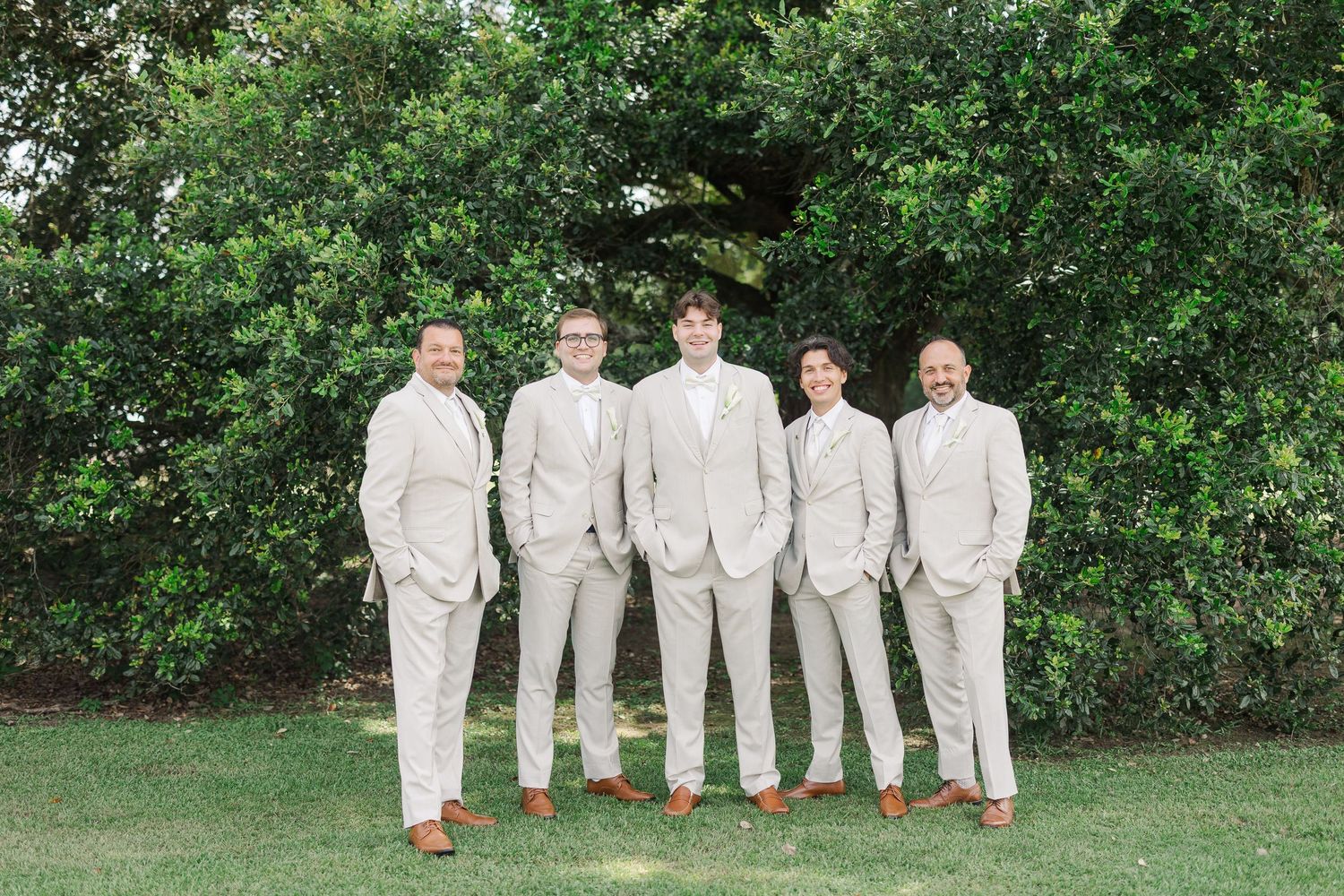 Five groomsmen in matching light beige suits with brown dress shoes pose outdoors in front of lush green trees.