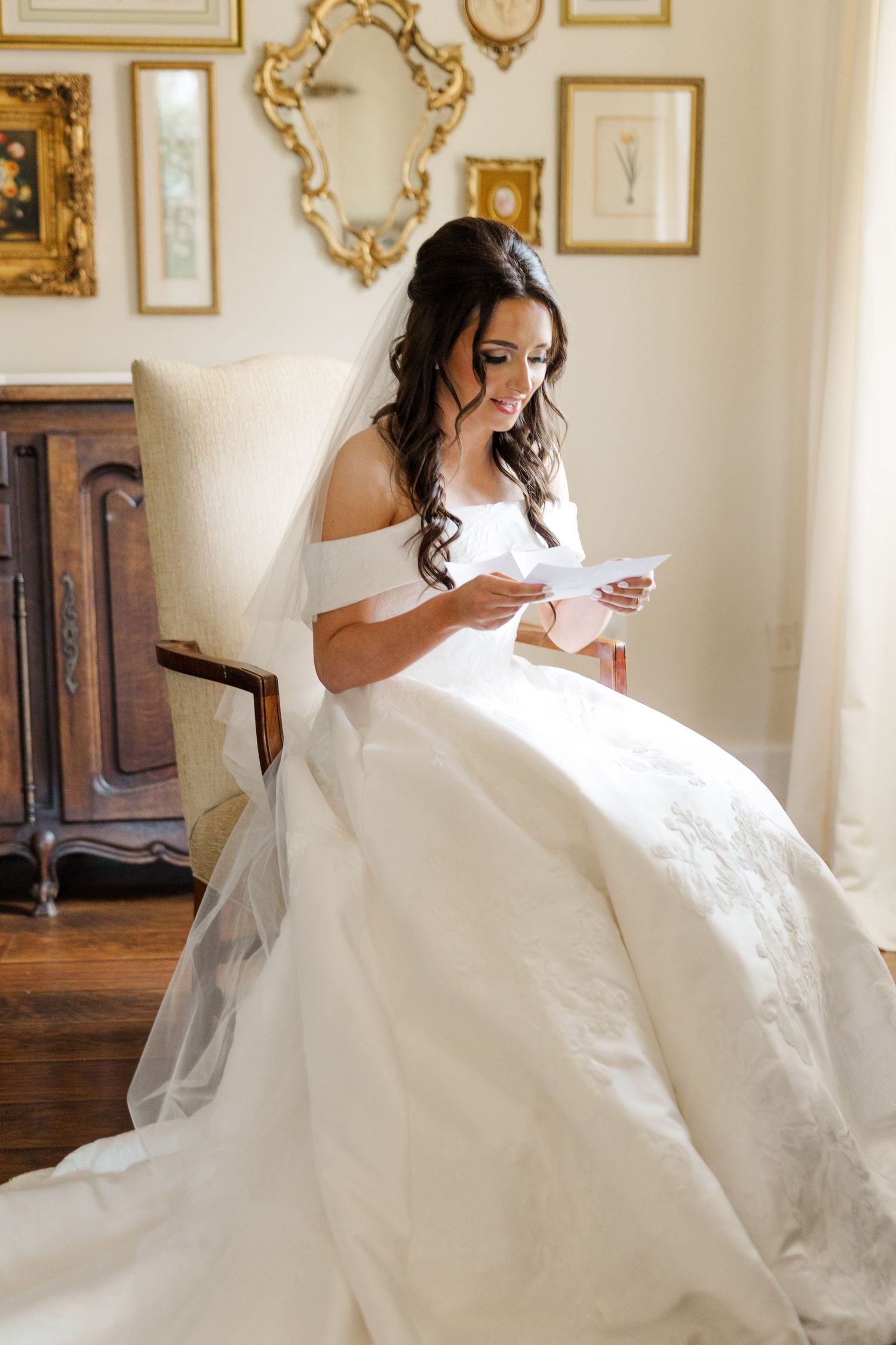 A bride in an elegant white off-shoulder gown reads a letter while sitting in an ornately decorated room with gold frames.