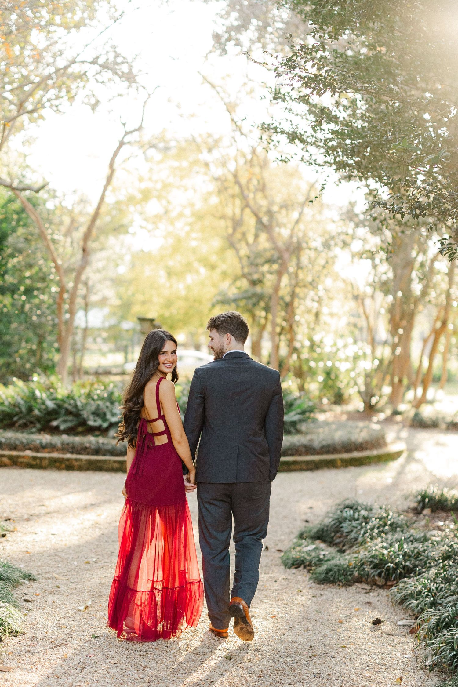 A couple walks hand in hand down a garden path wearing formal attire.