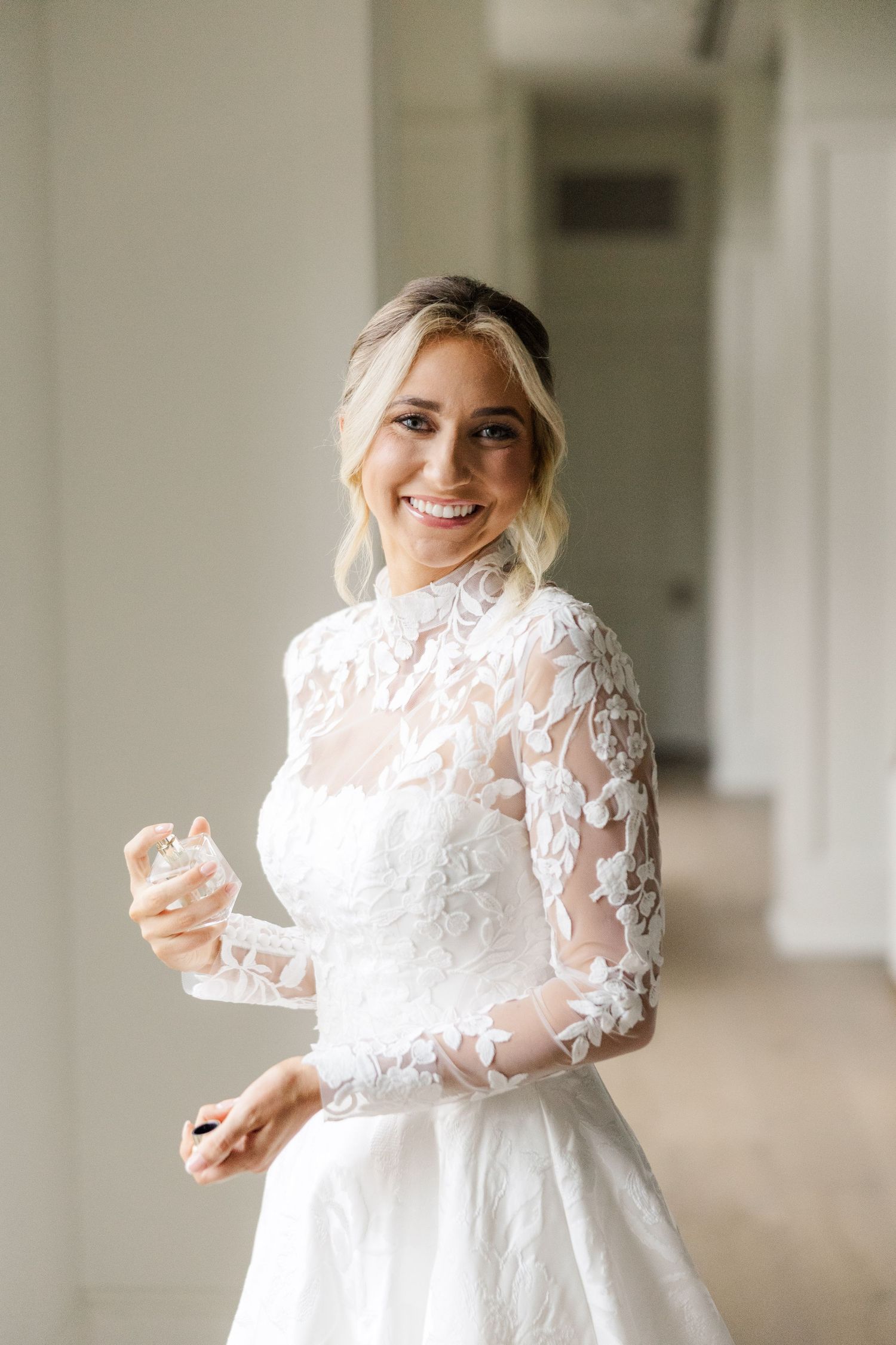 Bride in white lace gown smiles radiantly in sunlit church hallway.