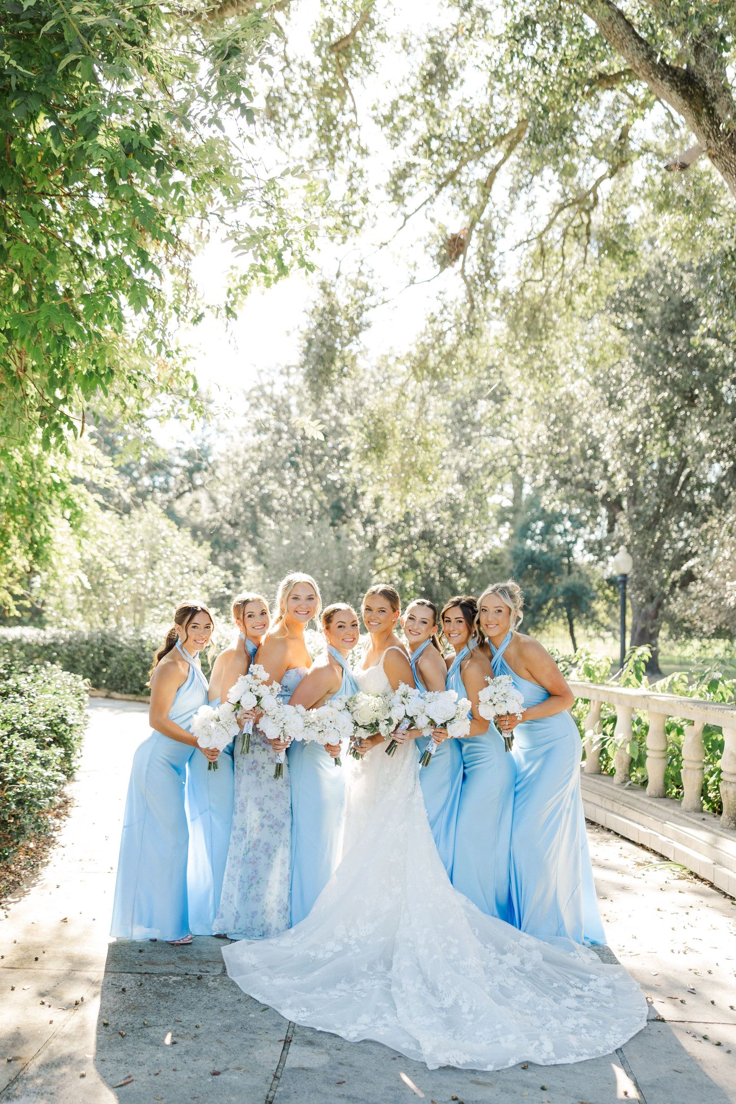 Bridal party in light blue dresses poses together outdoors under sunlit trees.