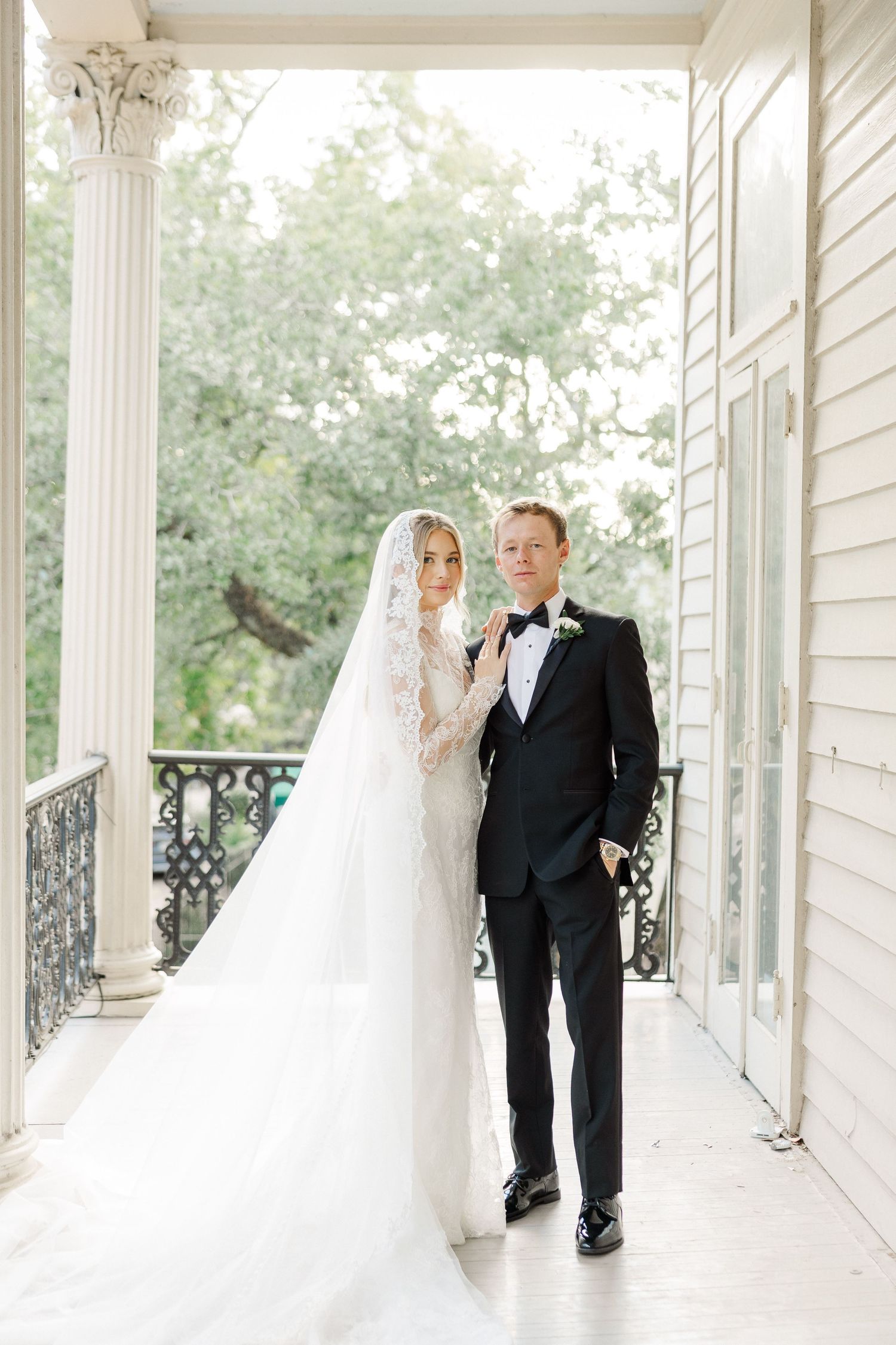 A wedding couple poses elegantly on a classic white columned porch with ornate railings.