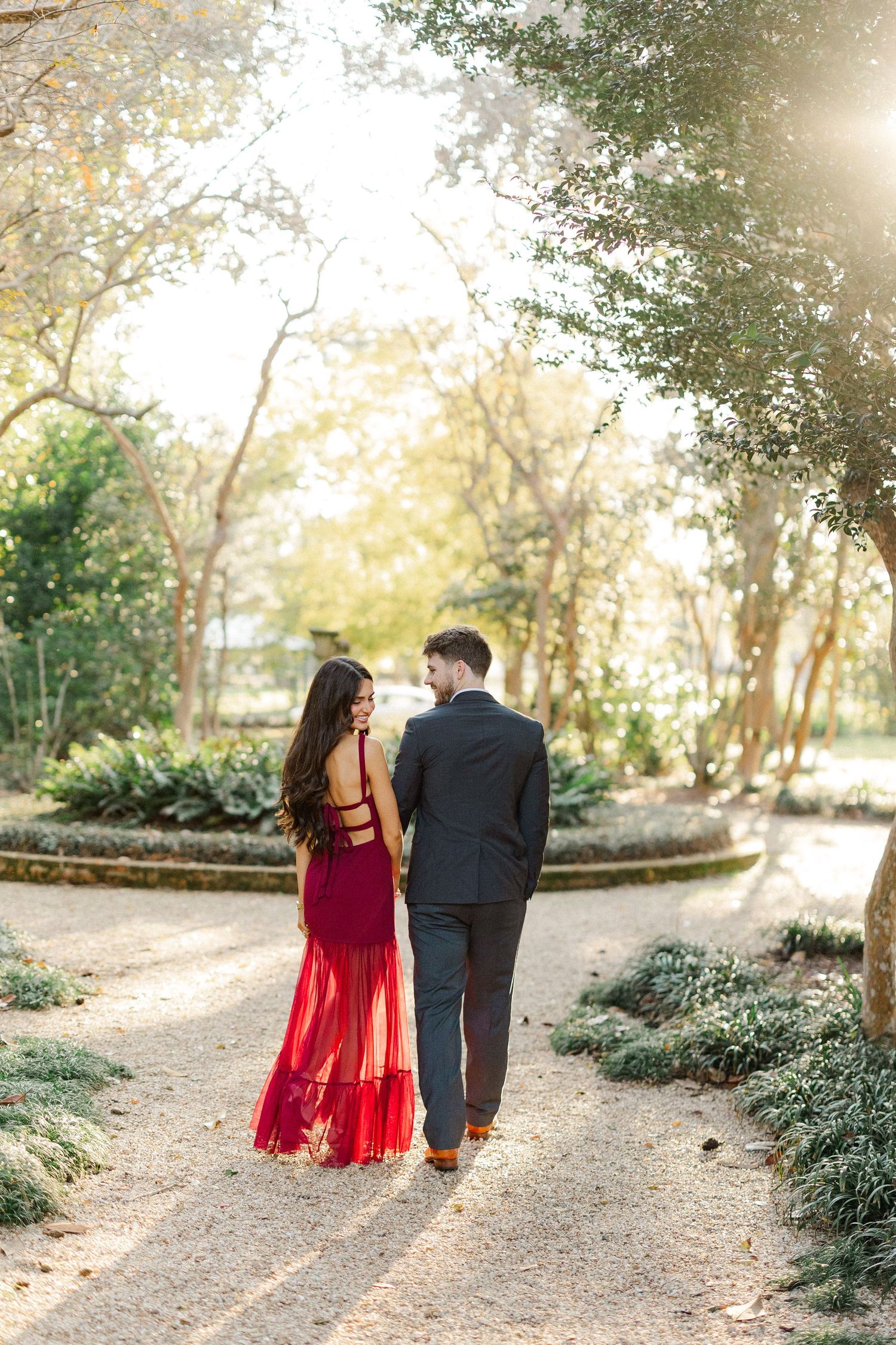 Couple walking hand in hand along a sunlit garden path wearing formal attire.