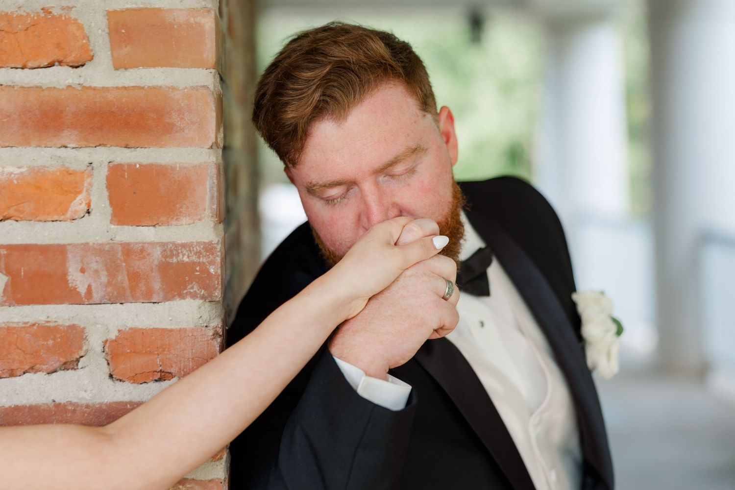A tender moment captured during a wedding as the groom kisses his partner's hand against a brick wall.
