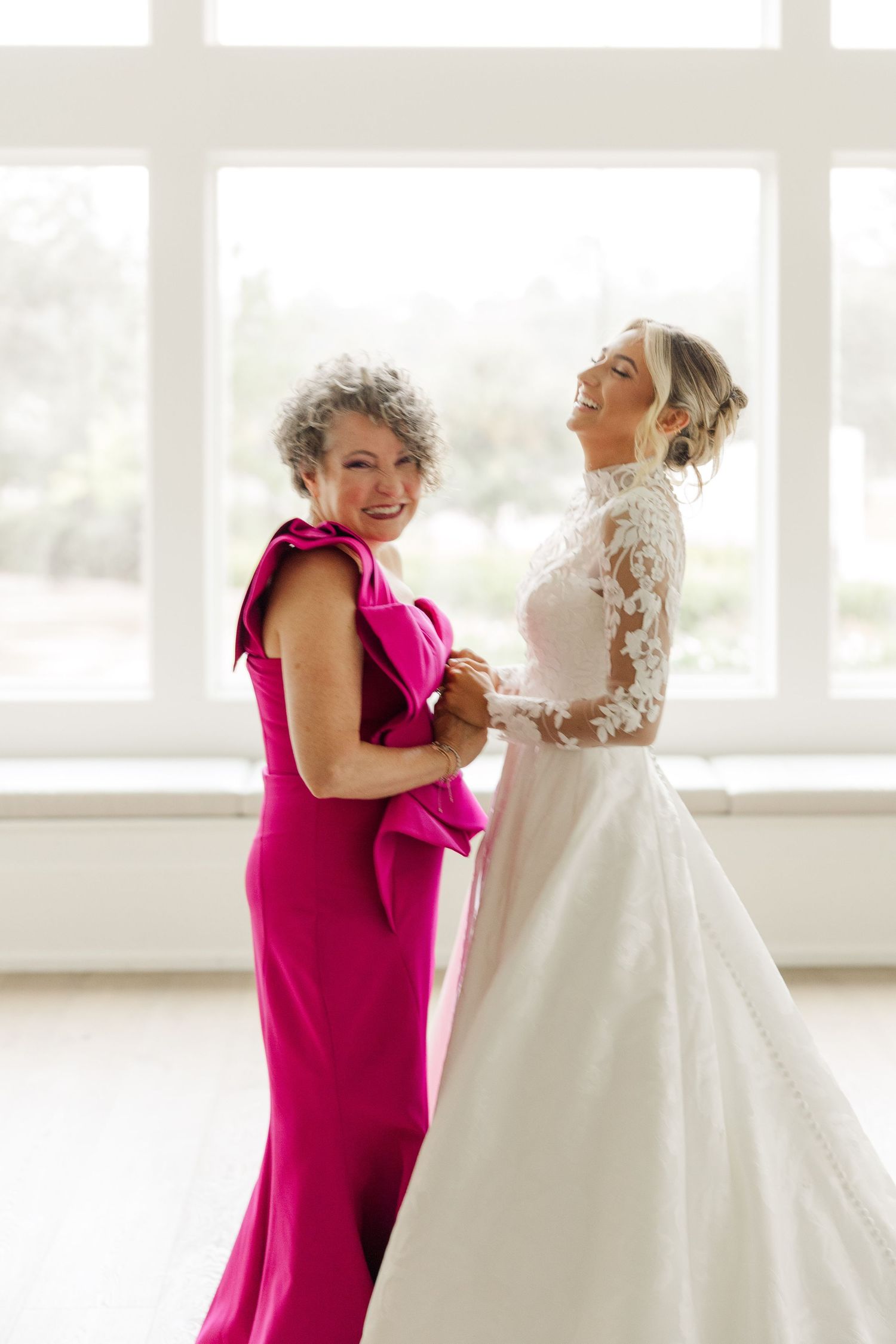 A bright pink bridesmaid dress contrasts with a white lace wedding gown being held up in a sunlit room.