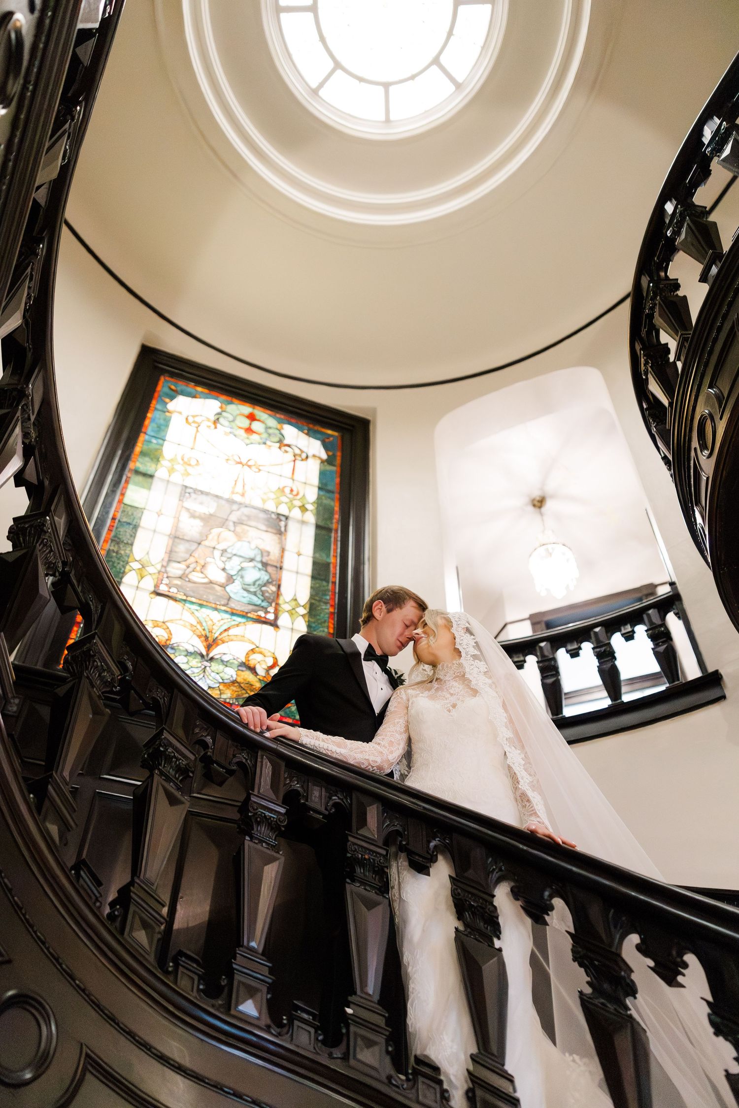 Elegant staircase wedding photo with dramatic circular window and ornate black railings.