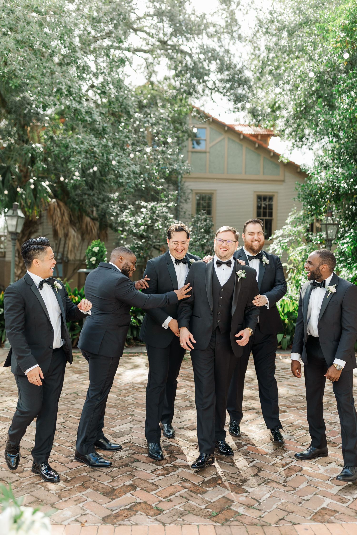 Wedding party groomsmen in black tuxedos share a candid moment laughing together in a garden courtyard setting.