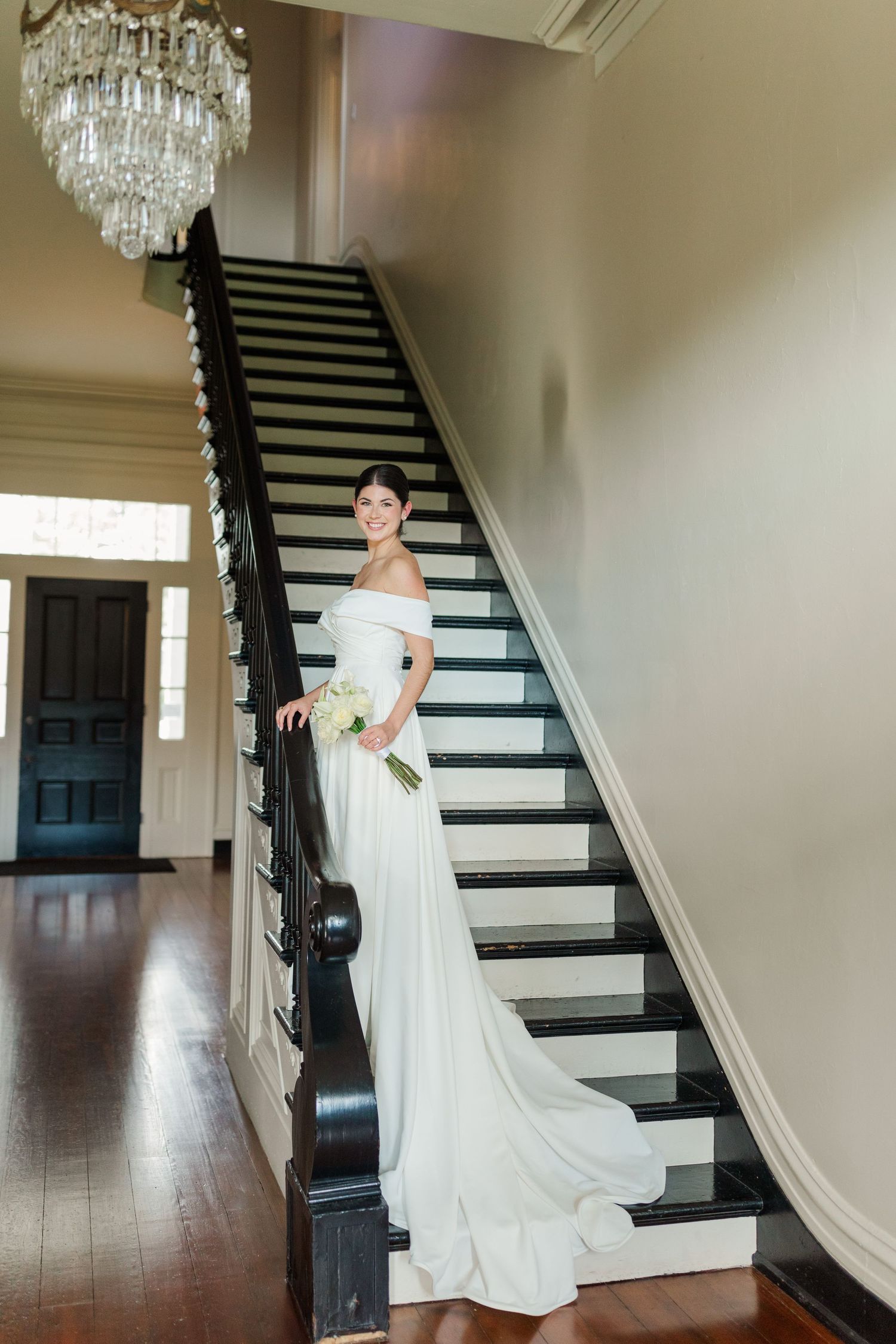 A bride in an off-shoulder wedding gown poses elegantly on a grand staircase beneath a crystal chandelier.