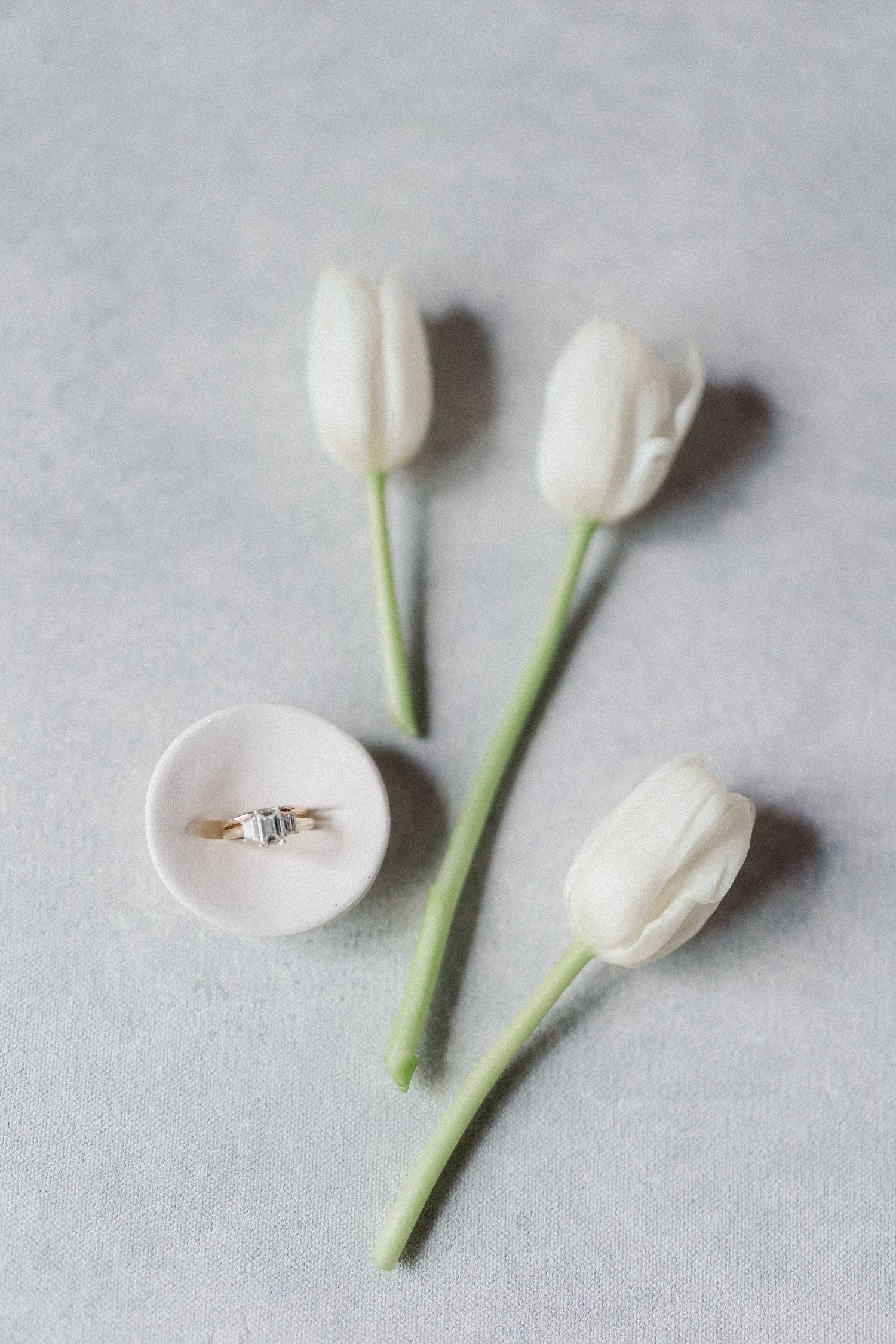 Gold diamond engagement ring in a white velvet box surrounded by three white tulips on a gray background.