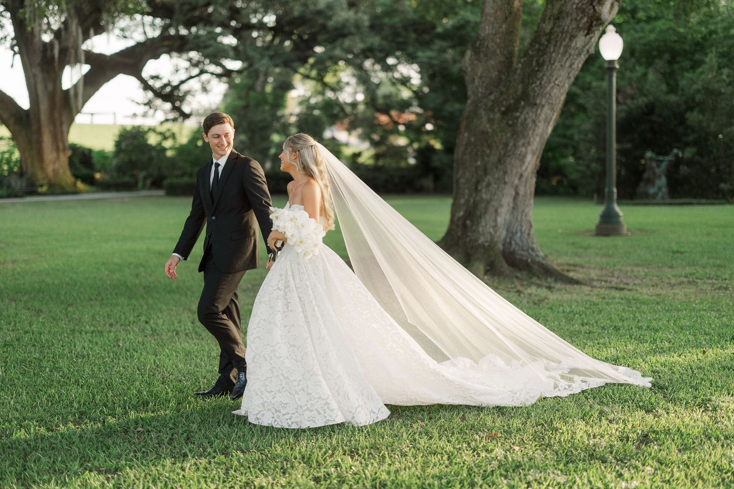 Bride in lace ballgown with flowing cathedral veil walks with groom across green lawn near oak tree.