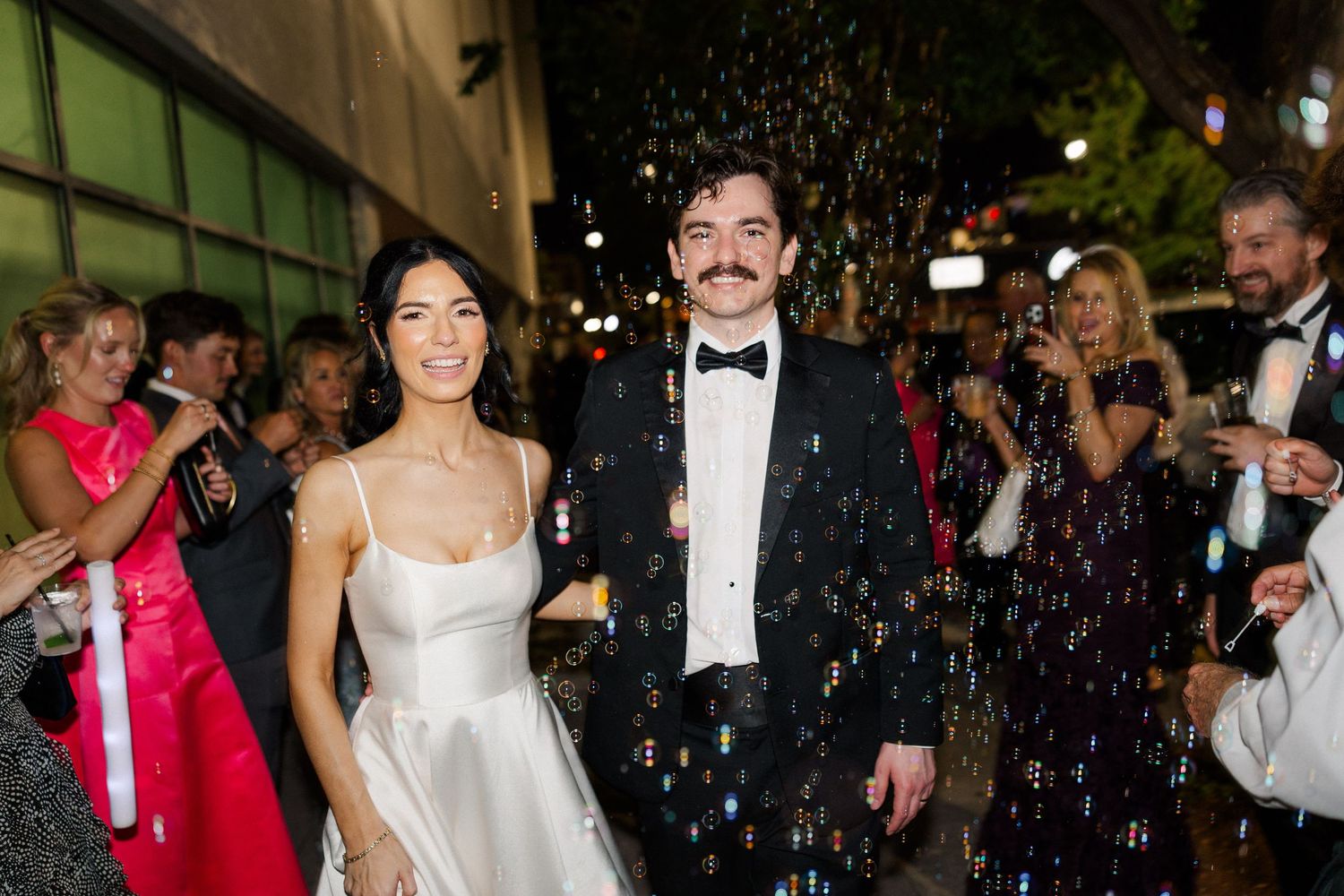 Bride and groom exit wedding venue through bubble send-off, bride in white satin gown, groom in black tuxedo.