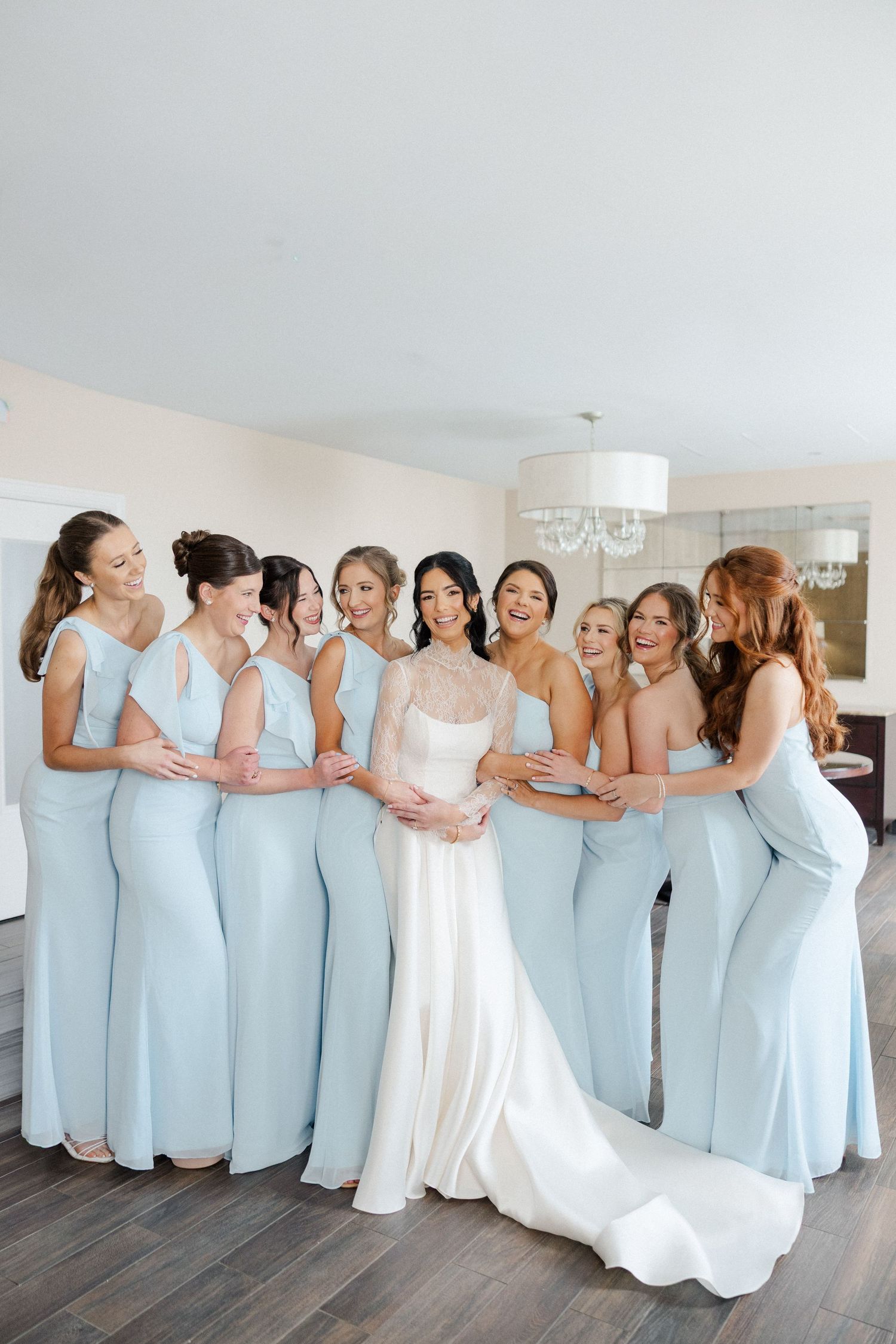 Bride in white gown surrounded by bridesmaids in light blue dresses laughing together indoors.