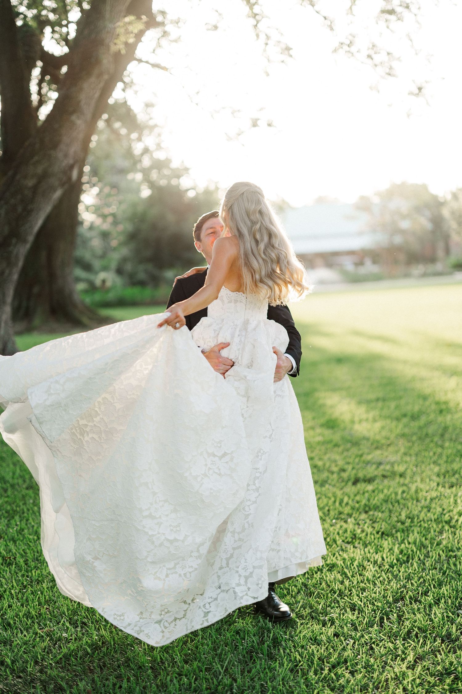 Groom lifts bride in stunning lace dress as her gown flows beautifully in outdoor wedding photo.