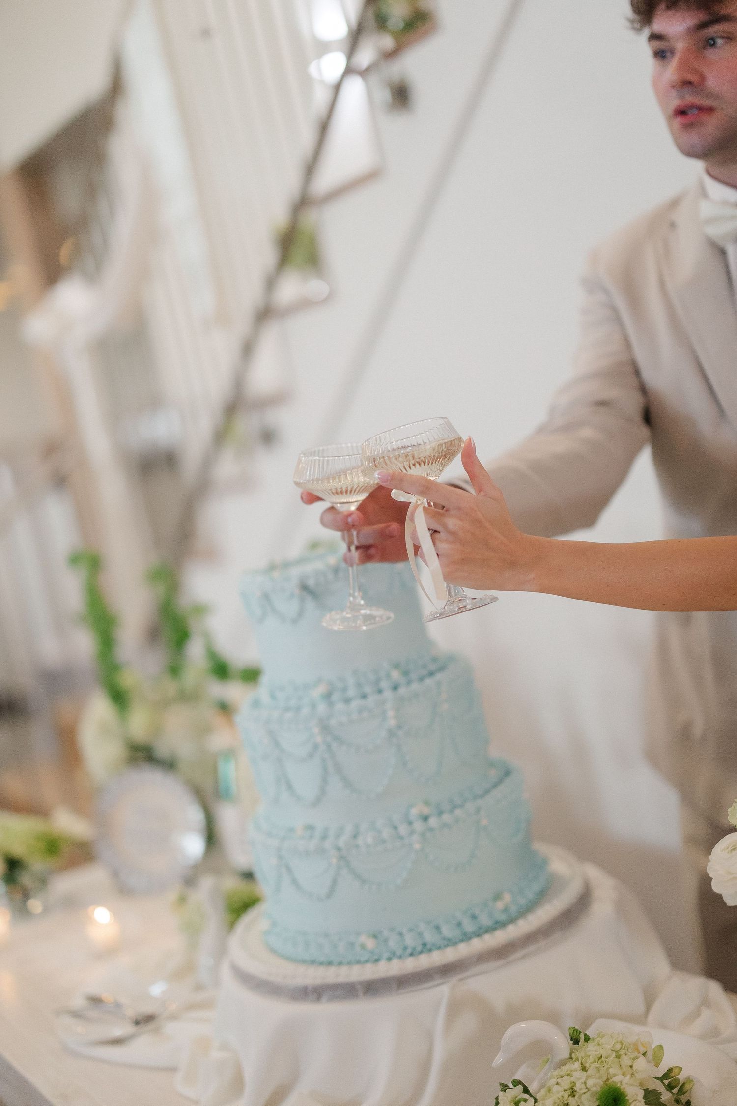 Couple toasting with champagne coupes beside elegant three-tier light blue wedding cake with pearl details.