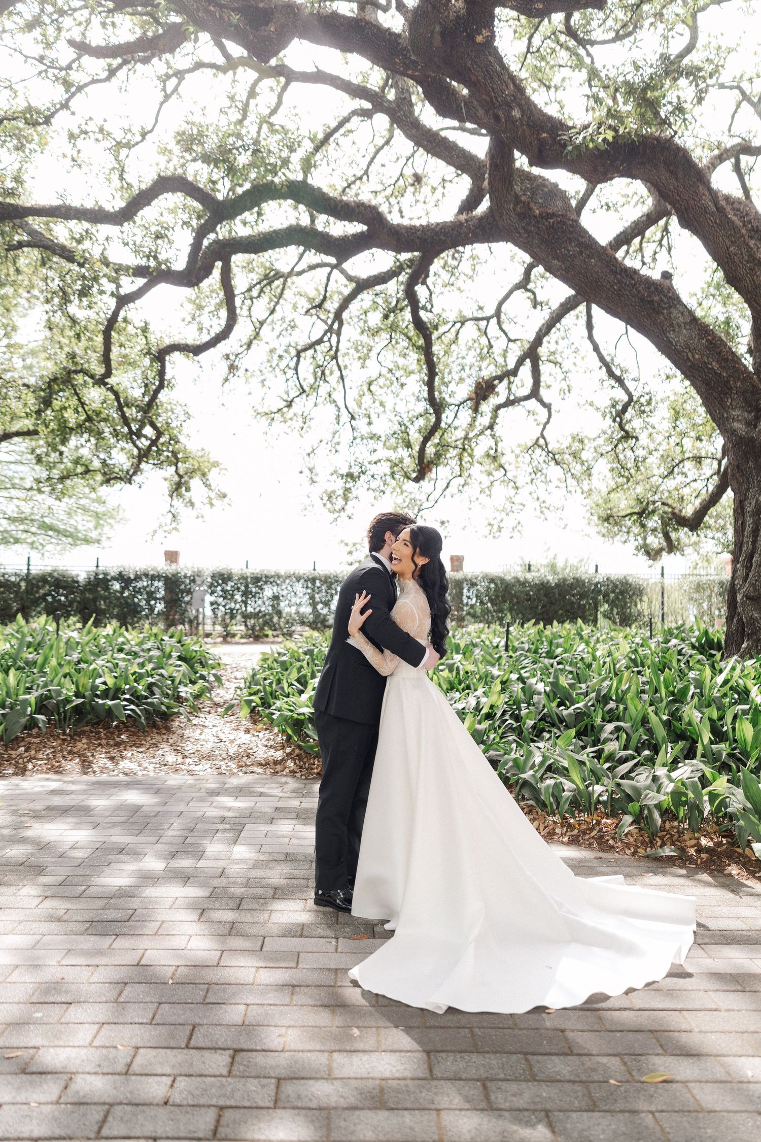Bride and groom sharing romantic kiss on garden pathway under large oak tree with lush greenery.