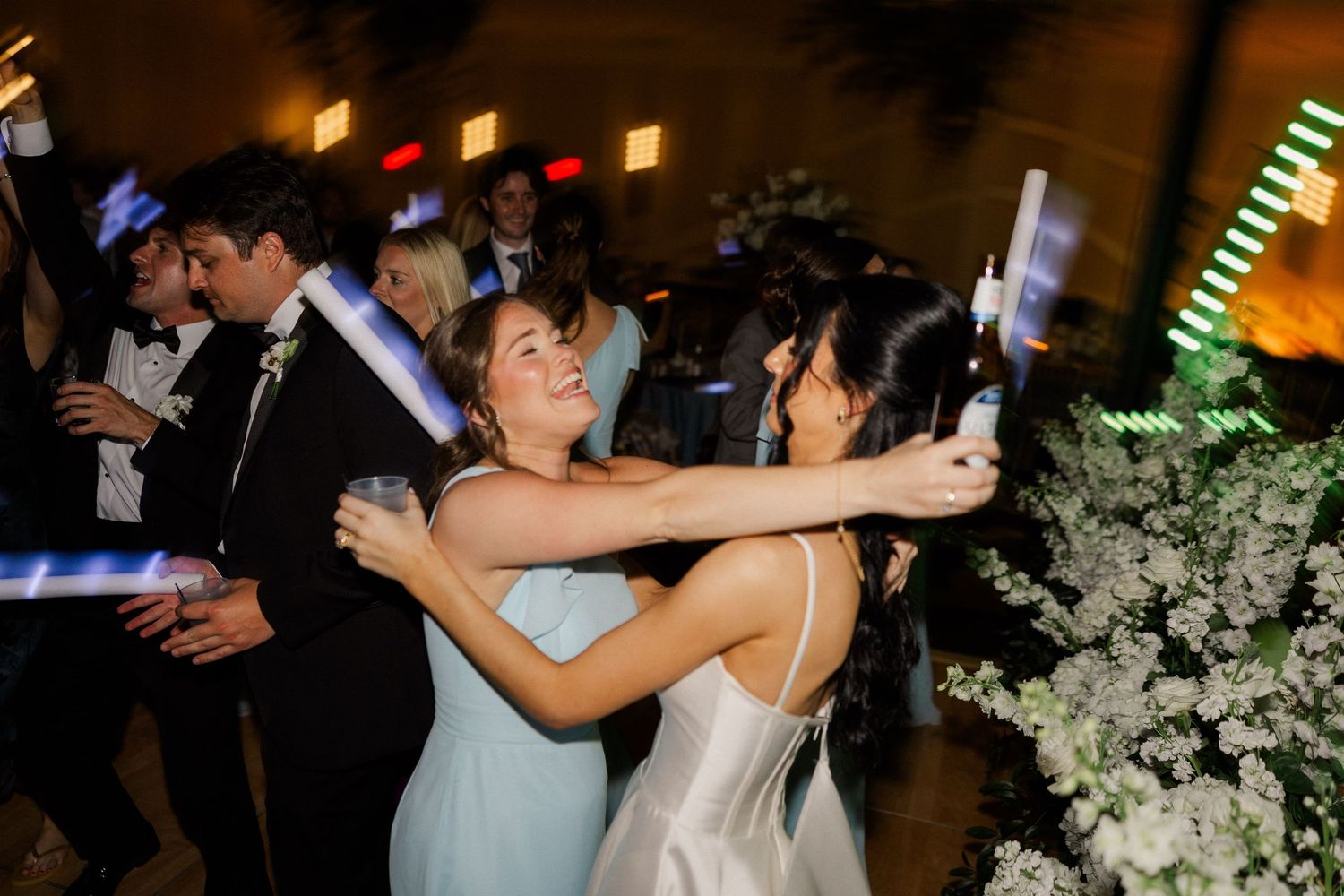 Bride and bridesmaid dance joyfully with glow sticks at lively wedding reception.