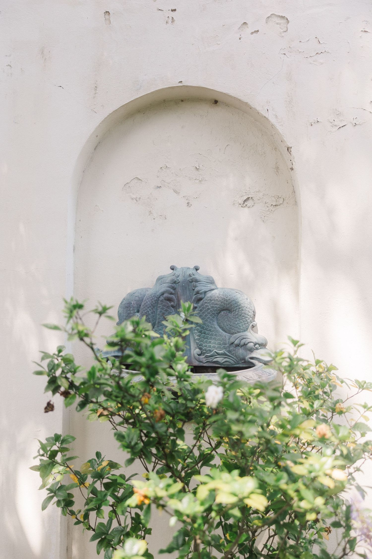 Stone dragon sculpture in arched wall niche surrounded by lush green foliage in a serene garden setting.