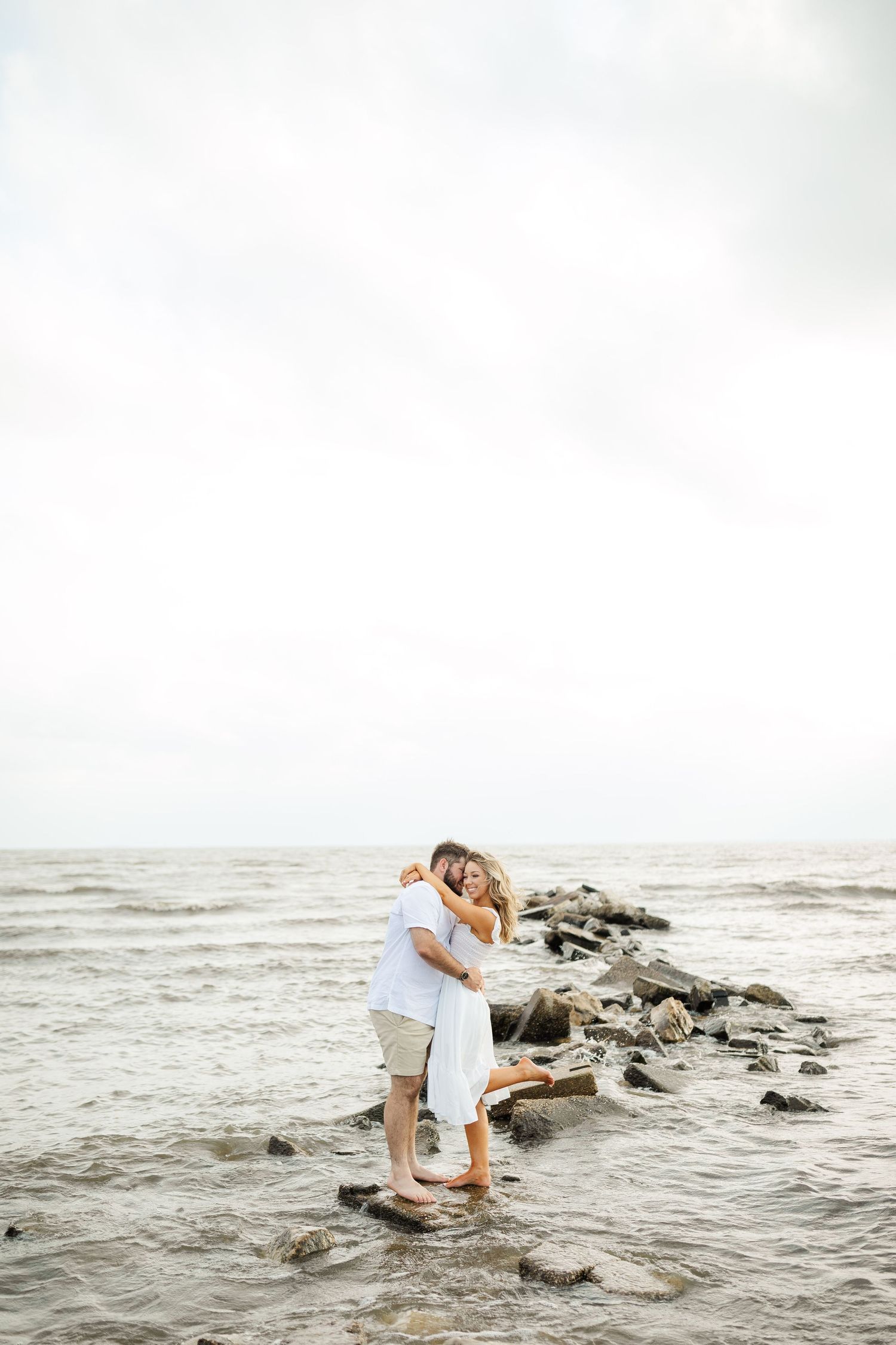 A romantic couple in white embraces while standing on rocky coastal shoreline at sunset.