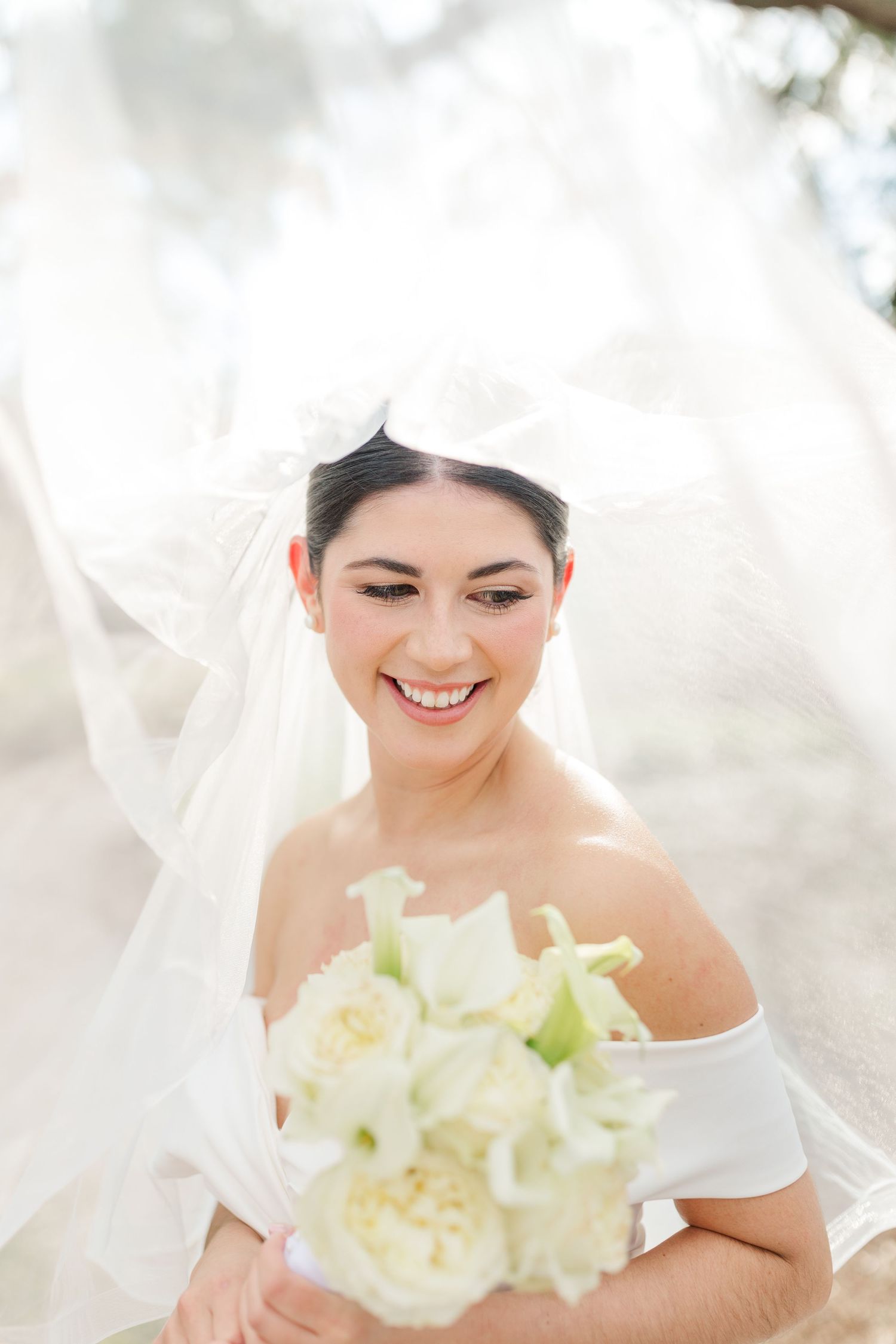 A radiant figure in wedding attire holds a delicate white and cream bouquet while wearing an off-shoulder gown and veil.