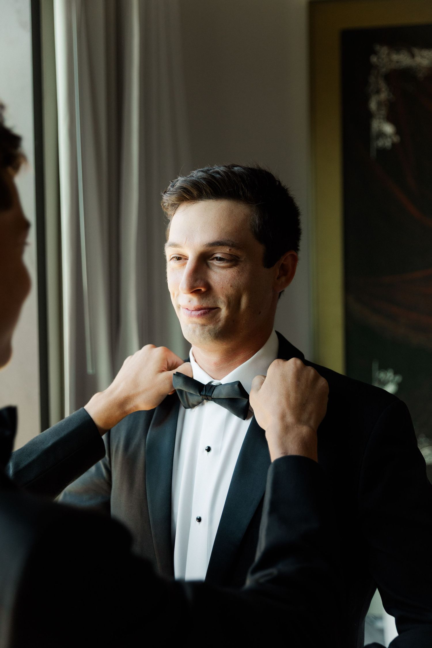 A groom in a classic black tuxedo and bowtie adjusts his collar while looking out a window.
