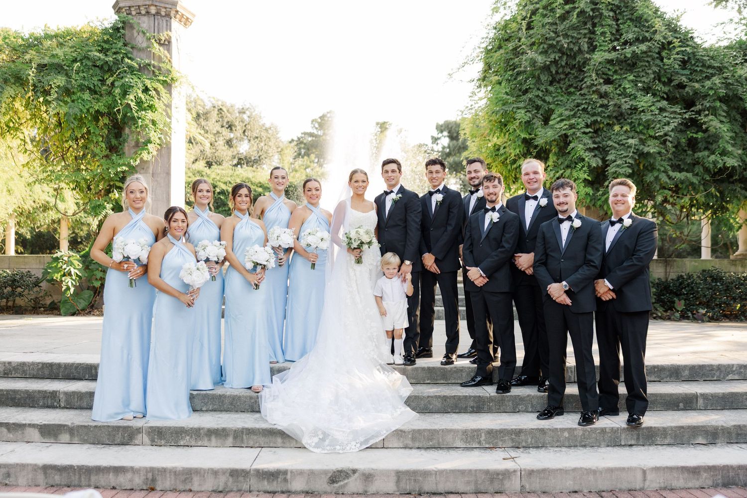 Wedding party in light blue bridesmaid dresses and black tuxedos pose elegantly on stone steps outdoors.